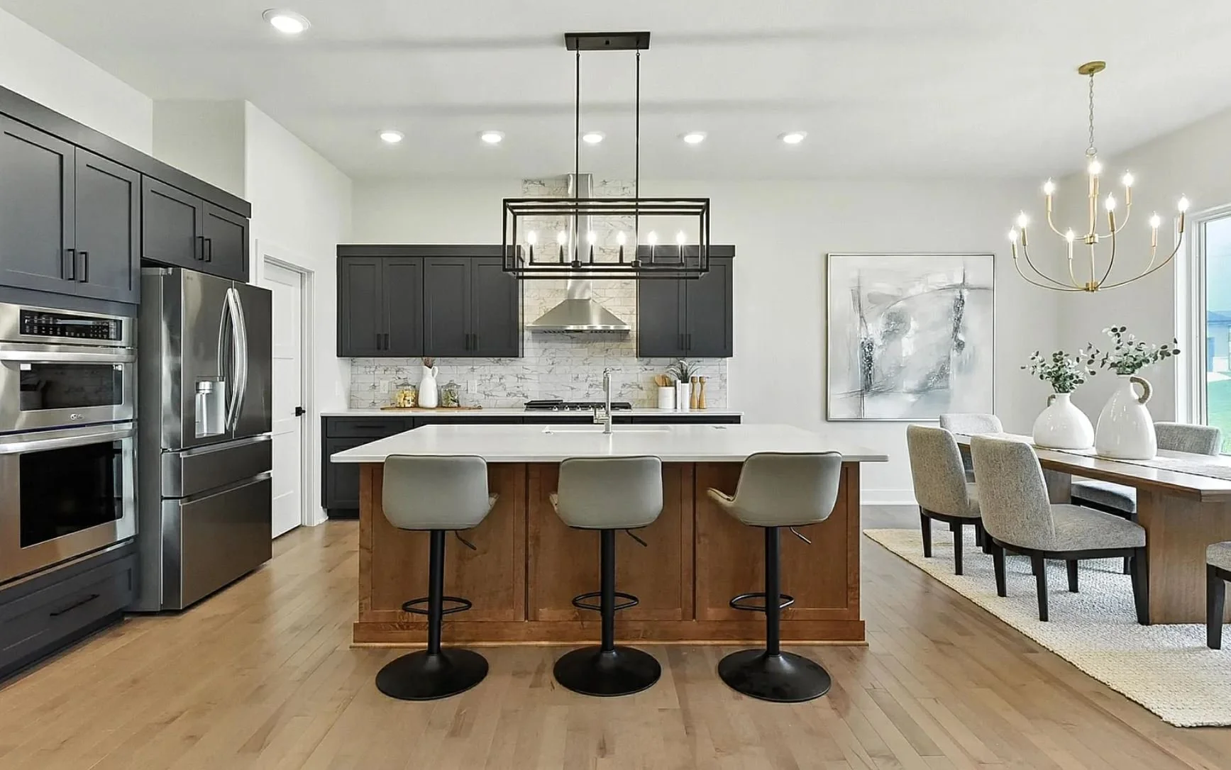 Modern kitchen with black cabinetry, stainless steel appliances, a white island with three beige barstools, light hardwood floors, and a dining area with a wooden table, beige chairs, and vases with greenery.