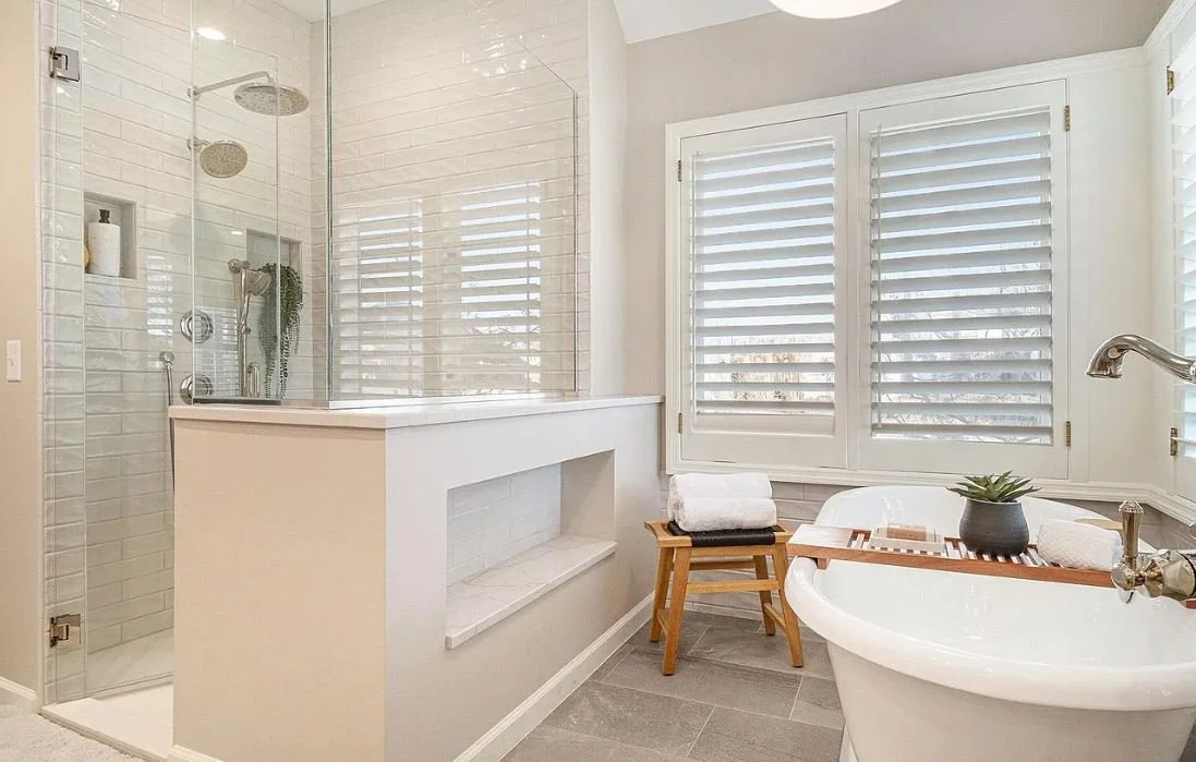 Modern bathroom featuring a walk-in shower with glass doors, a clawfoot bathtub with a wooden tray and towels, large window with white blinds, and neutral-toned decor.