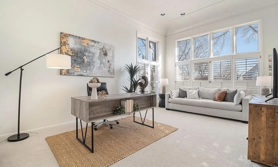Living room with white walls, large windows with shutters, a white sofa, a wooden desk, a black desk chair, a tall plant, and a floor lamp.