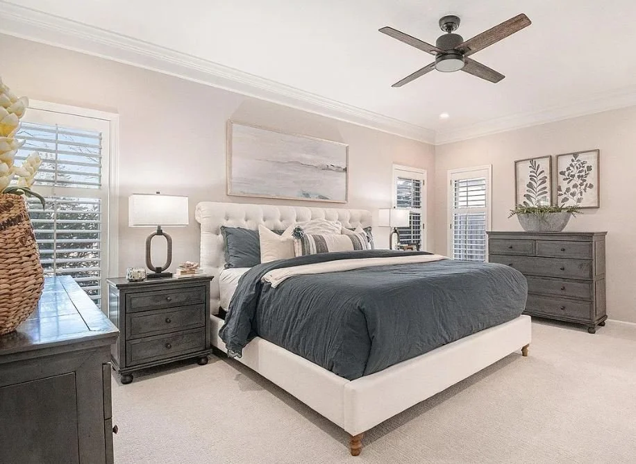 A bedroom with a large bed, dark gray bedding, and a tufted white headboard. Two nightstands with lamps, a dresser with a plant, and framed botanical art on the walls. Windows with white shutters and a ceiling fan.