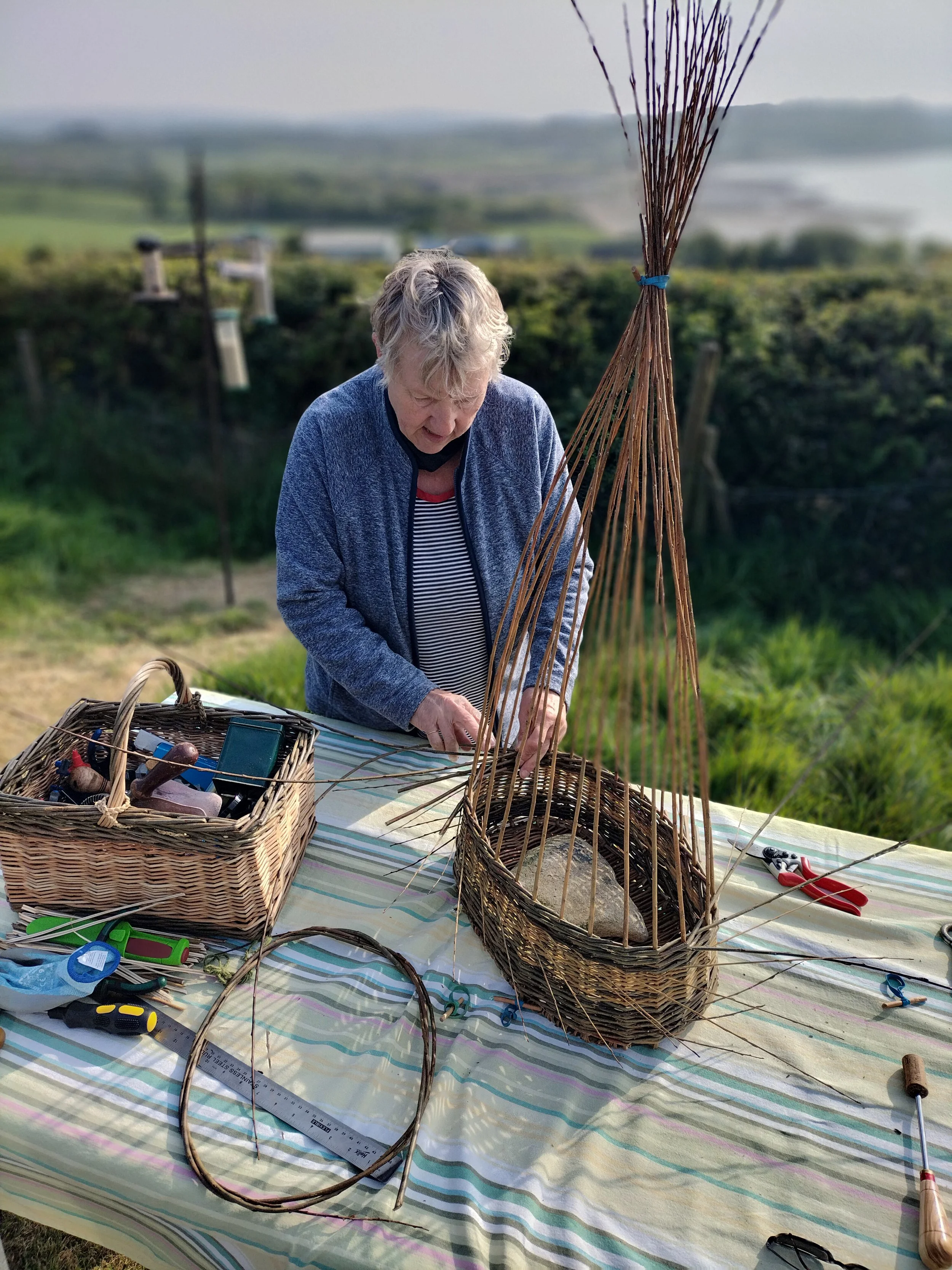 Angela looking down and weaving willow at a table with tools and baskets around her. The background is of rolling hills and the sea.