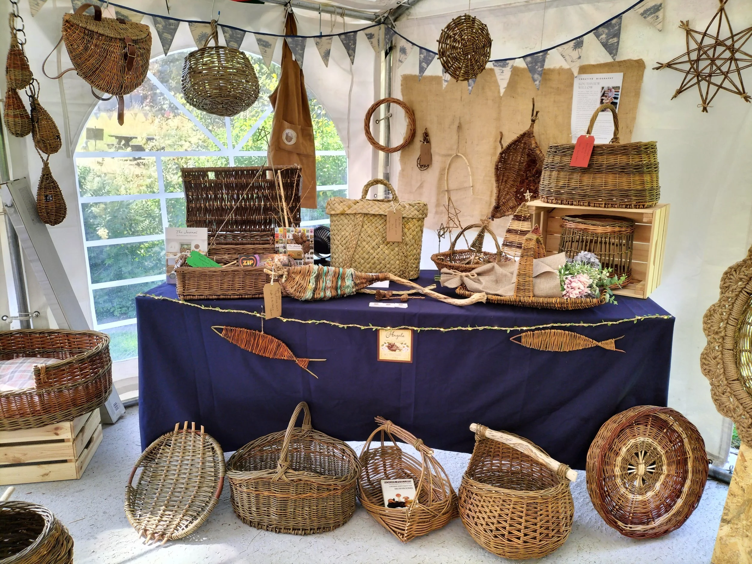 A display of woven baskets and decorative straw art at an outdoor market stall, with a blue tablecloth and a backdrop of banners and hanging straw decorations.
