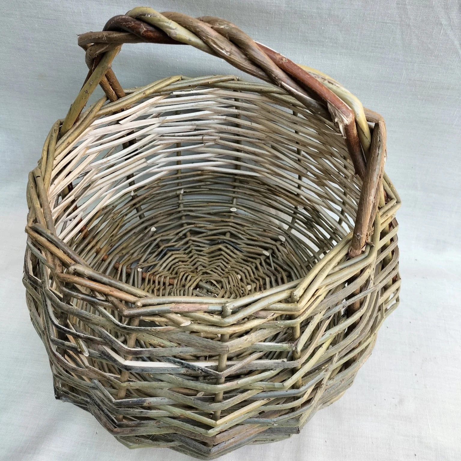 A woven wicker basket with a rounded shape, open top, and a handle at the top, made from natural twigs and branches.