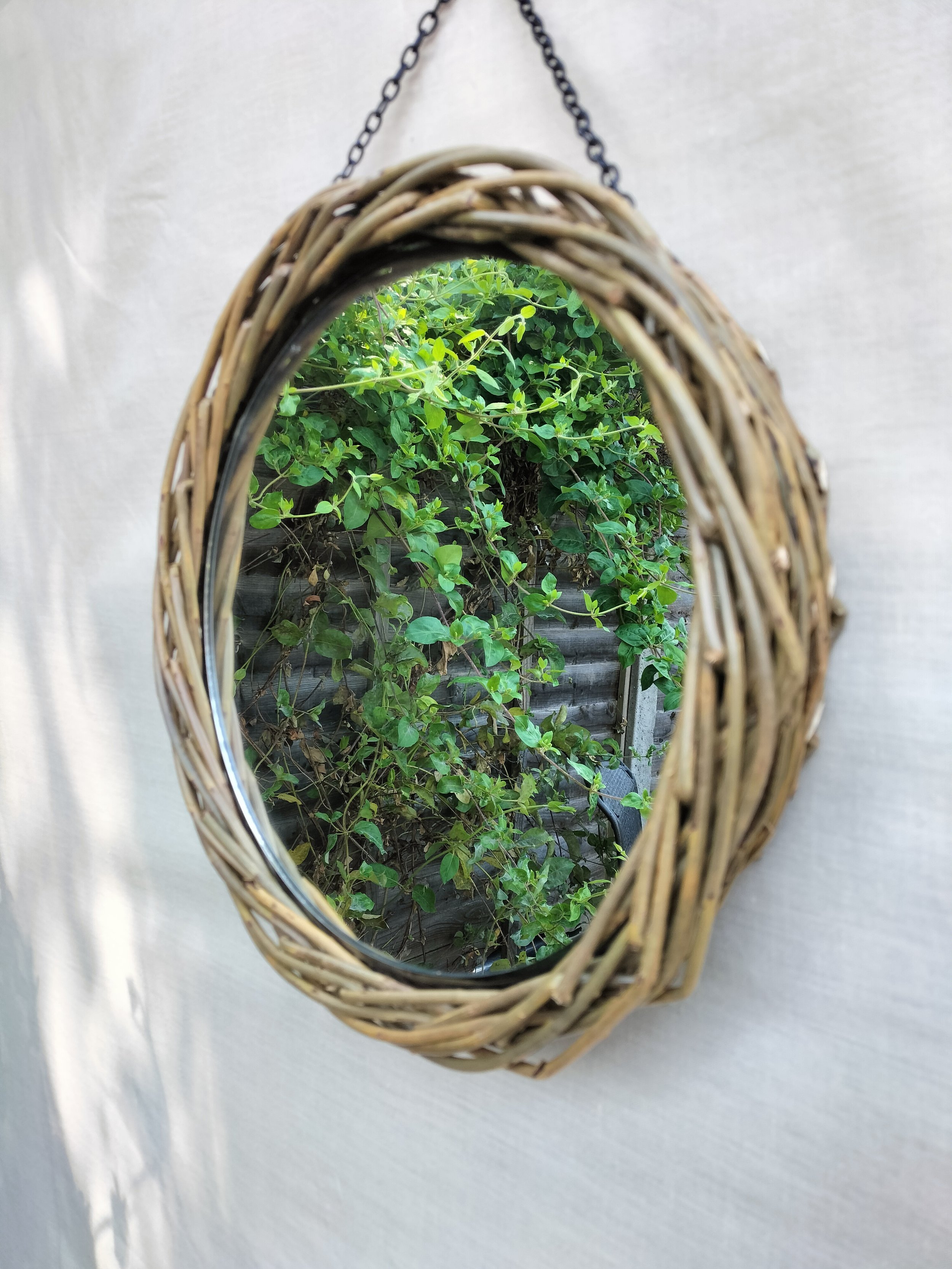 Oval mirror with a woven wicker frame hanging on a wall, reflecting green leafy plants and wooden fencing.
