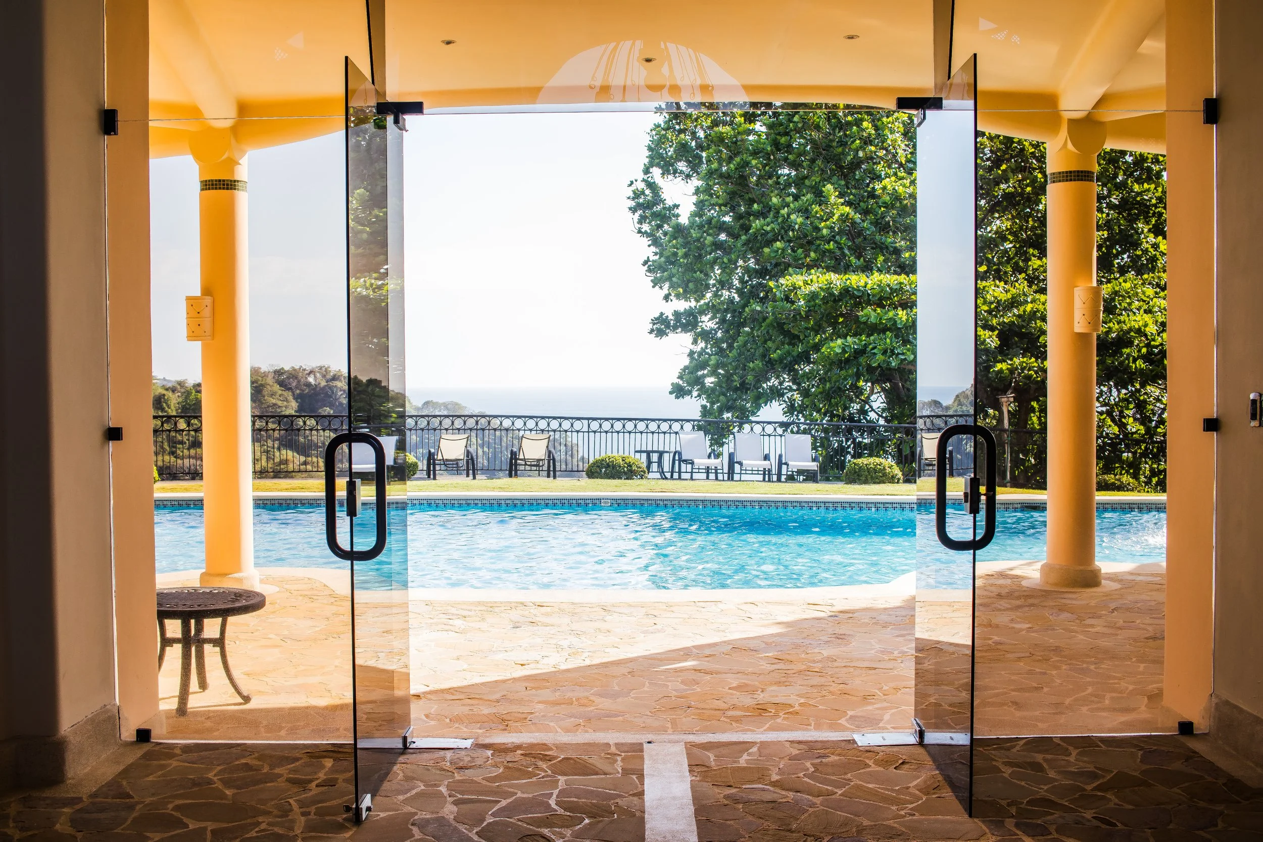 Vista de una piscina desde el interior de un espacio con puertas de cristal abiertas, rodeada de sillas y árboles, con un cielo despejado y vistas de la naturaleza en el fondo.