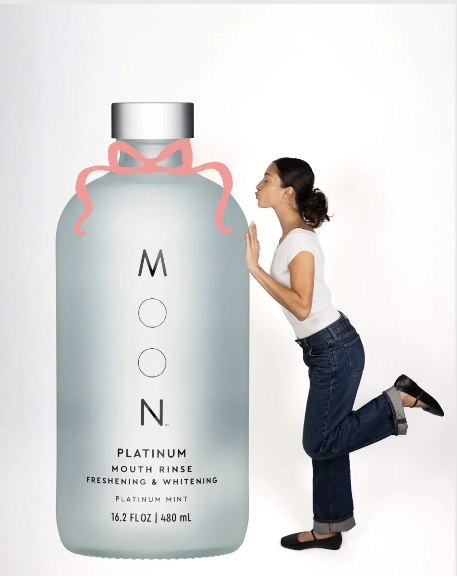 A woman kneeling and leaning towards a large bottle of mouth rinse, with her eyes closed, about to kiss the bottle's cap, which is decorated with a pink ribbon.