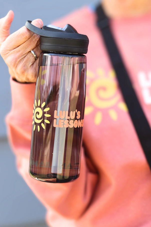 A clear tumbler with a black lid, labeled "LULU'S LESSONS," being held by a person wearing a pink sweatshirt with a yellow sun logo.