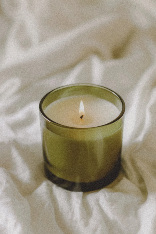 A lit white candle in a green glass holder placed on a crumpled white fabric surface.