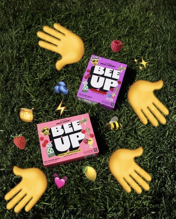 Two boxes of BE UP sour gummy candies and scattered emoji-style graphics of hands, berries, a heart, a bee, a lightning bolt, and other icons on grass.