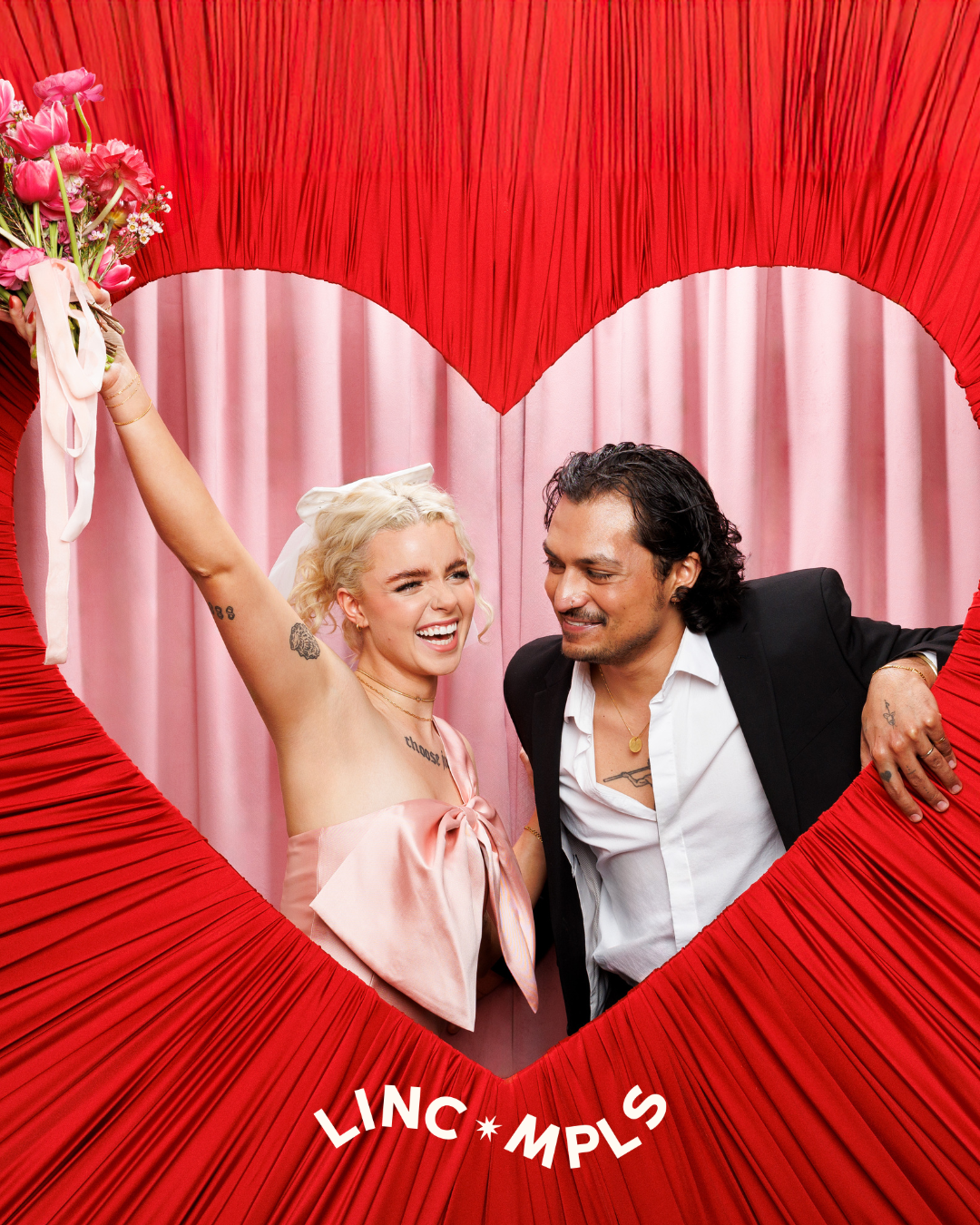 A smiling woman holding a bouquet of pink flowers and a man smiling, both behind a large red heart-shaped decoration with pink curtains in the background, with the words 'LINC * MPLS' at the bottom.