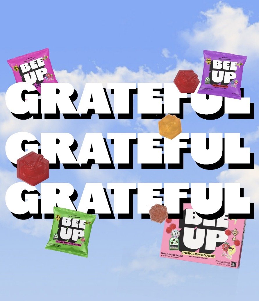 Graphic featuring the words "GRATEFUL" repeated three times in bold white text with black shadows, set against a background of blue sky and clouds. Scattered around are colorful gummy candies and packets of BEAR UP fruit snacks in various flavors inc