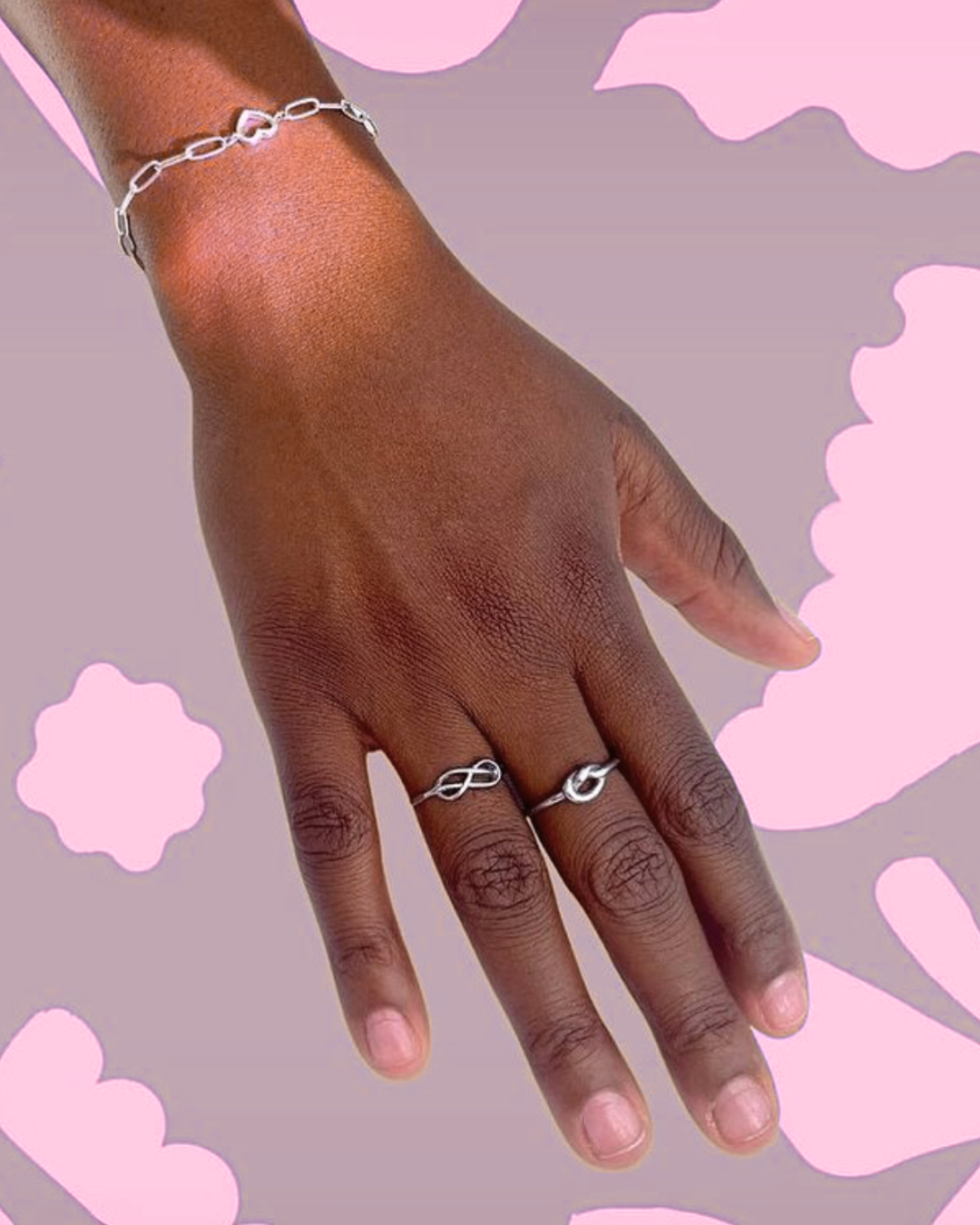 A hand with dark skin wearing a silver infinity bracelet and two silver rings with infinity and wave designs, against a background of pink cloud shapes.
