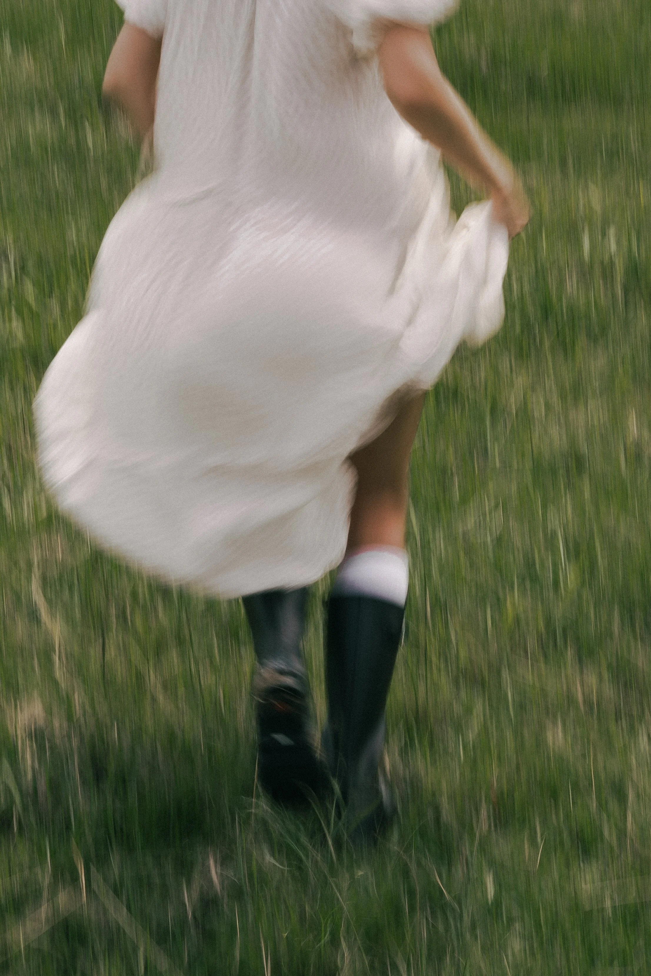 A person running in a white dress and black rain boots on green grass.