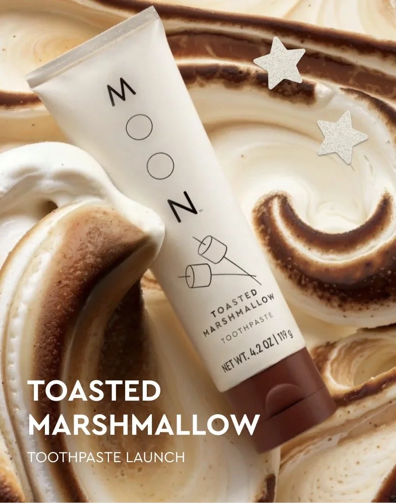 A tube of toasted marshmallow flavored toothpaste lying on a swirl of marshmallow and chocolate-colored cream, with two glittery silver star stickers nearby.
