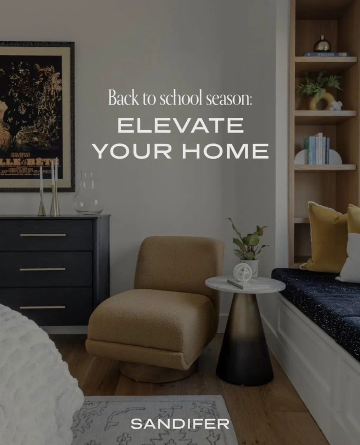 Living room with beige armchair, black dresser with decor, small white round side table with plant and remote, built-in wooden bookshelf with plants and books, bed with yellow and white pillows, framed artwork on wall, and a rug.