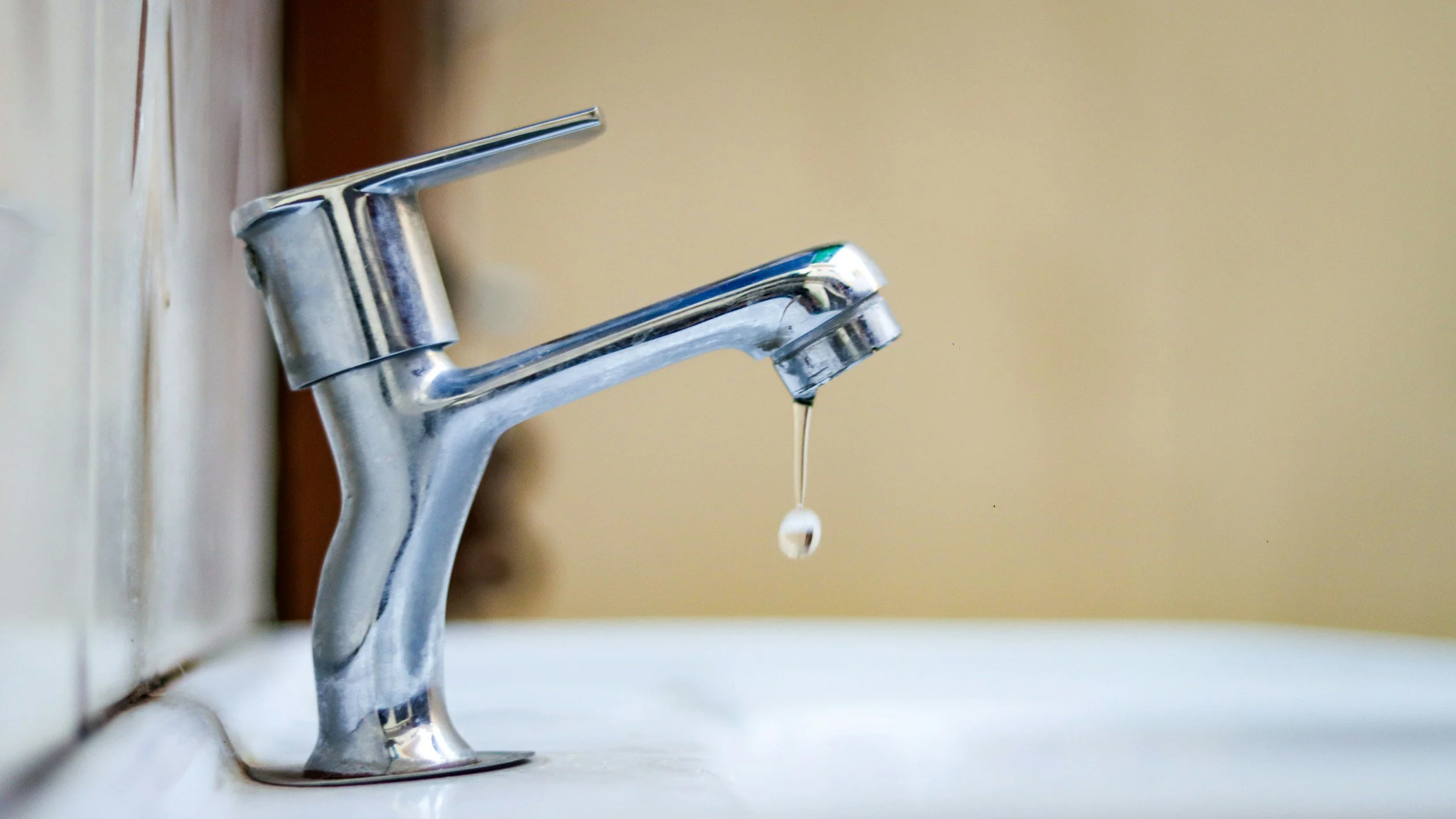 Close-up of a silver faucet with a droplet of water hanging from the spout over a white sink.