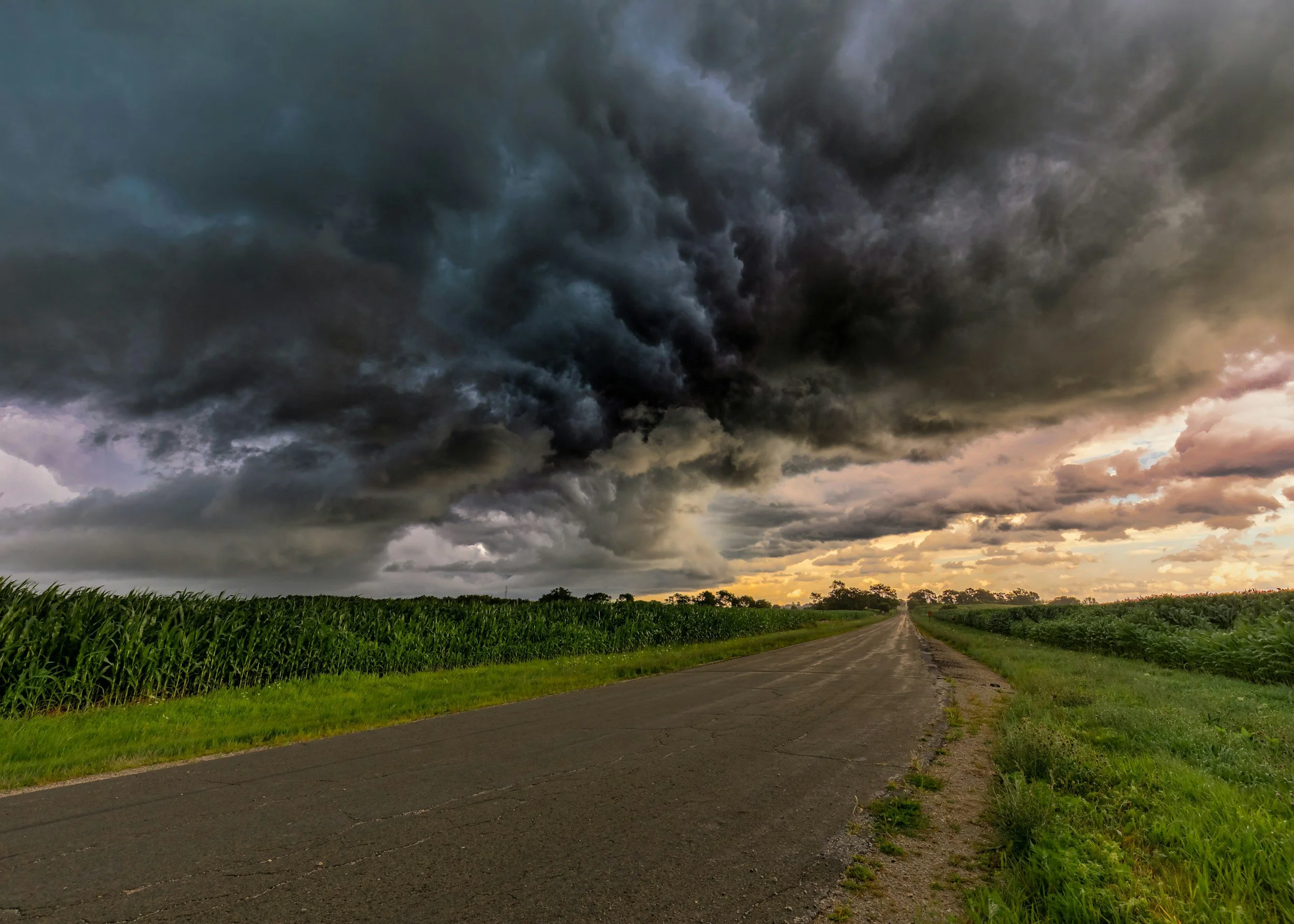 Dark storm clouds over a rural road bordered by green fields, with a distant horizon and an unsettled sky.