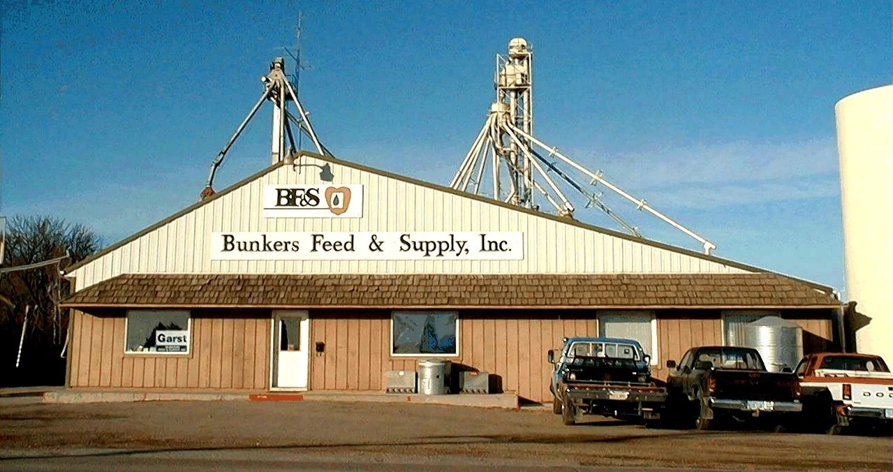 A beige building with a brown shingle roof and a sign that reads "Bunkers Feed & Supply, Inc." in black letters. There are three cars parked in front of it and a large grain silo structure behind.