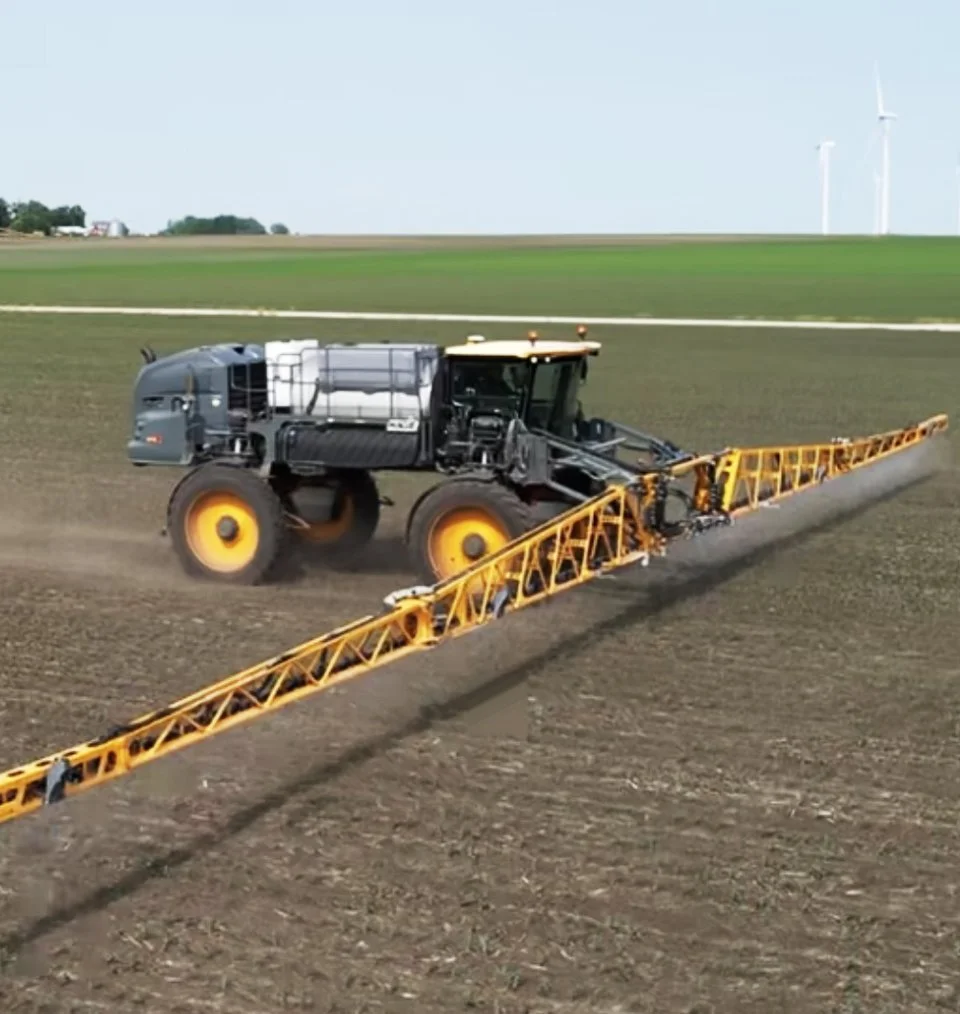 A large yellow and black tractor is spraying fertilizer or pesticides on a brown agricultural field with wind turbines in the background.