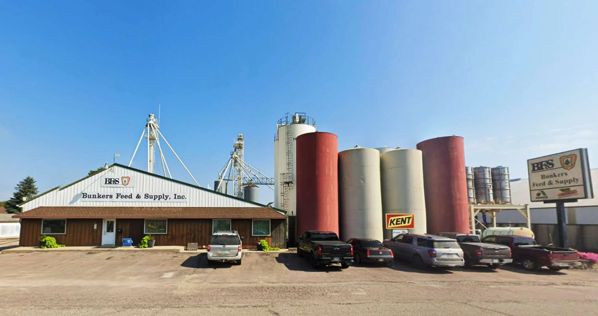 Bunkers Feed & Supply, Inc., with several large colored storage silos, parked cars in front, and a clear blue sky.