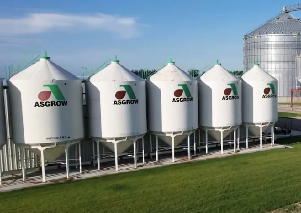 Six white grain silos with the ASGROW logo and text, standing on a grassy area with a blue sky in the background.
