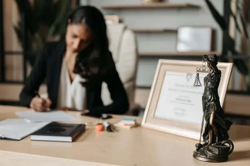 A woman sitting at a desk with legal documents, a framed certificate, and a Justice of the Peace statue.