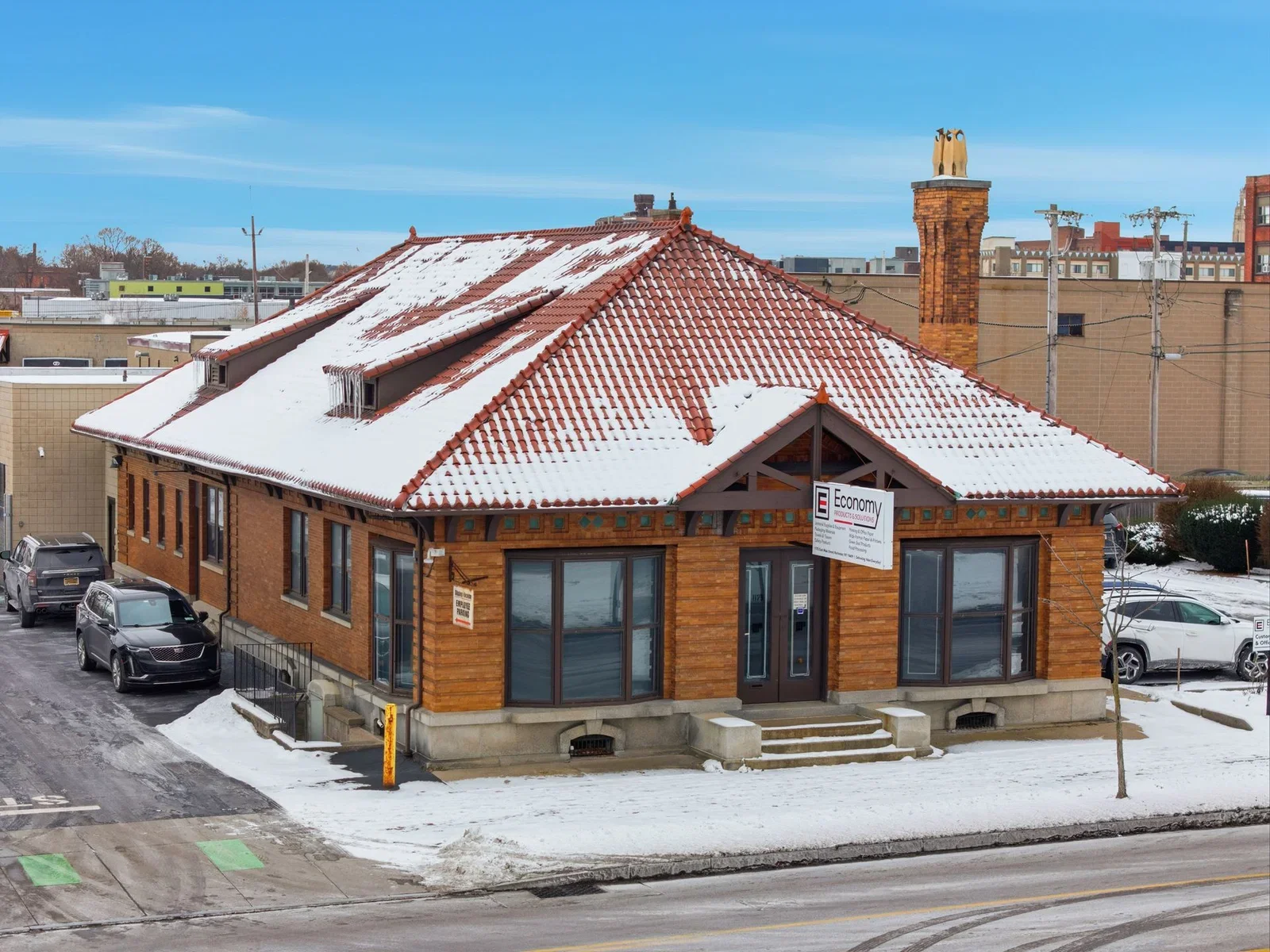 A building with a snow-covered red tile roof and brick exterior, with cars parked outside on a snowy street.
