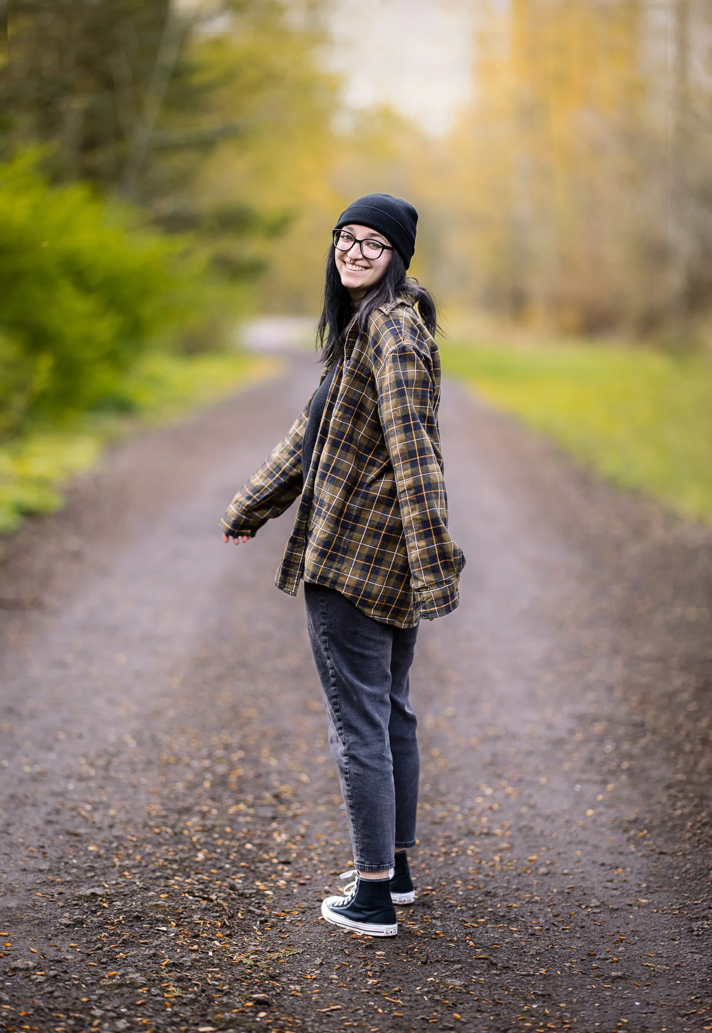 A young woman with glasses, wearing a black beanie, plaid jacket, and black sneakers, stands on a dirt path surrounded by autumn trees, smiling at the camera.