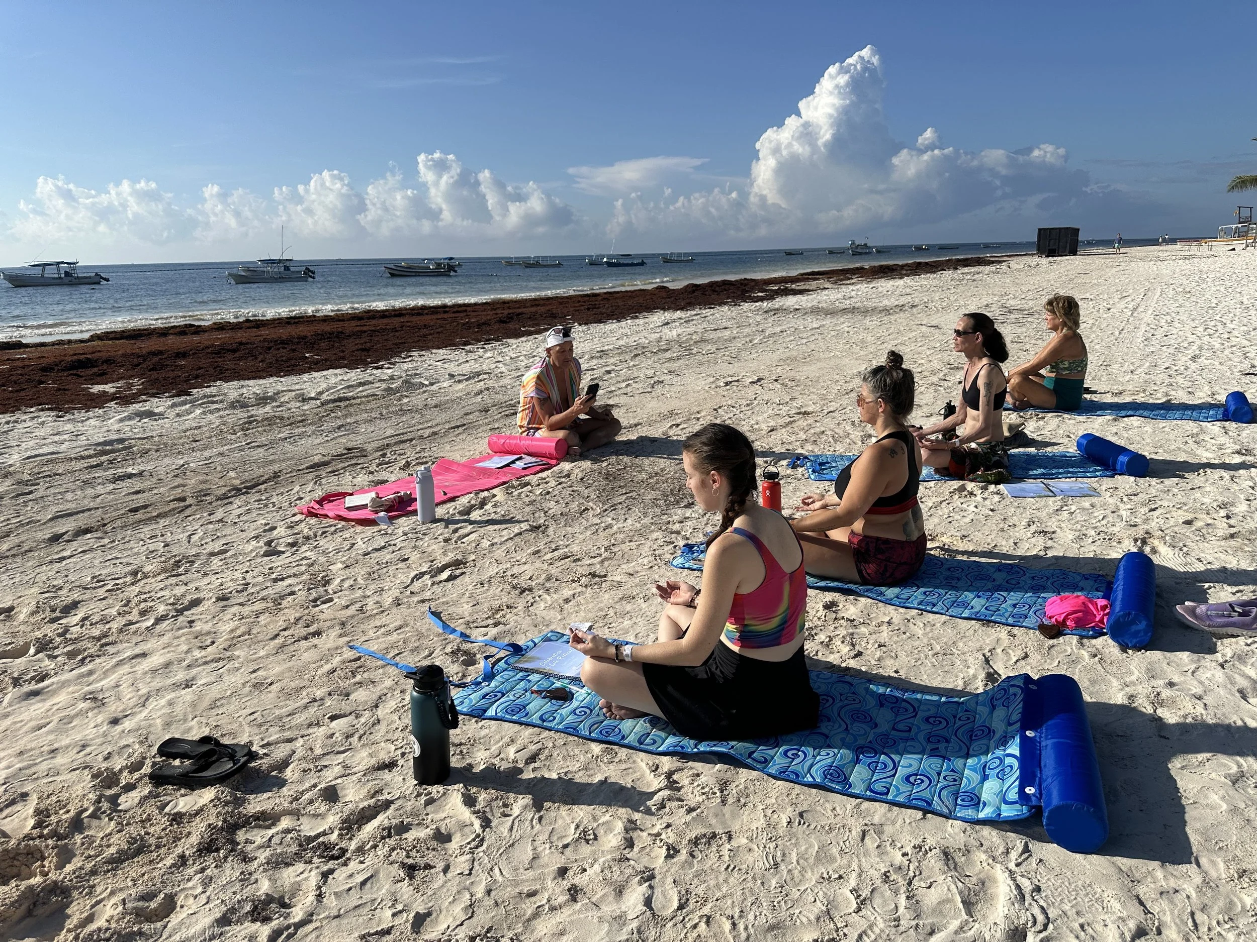 Group of women participating in a yoga class on a sandy beach with boats anchored offshore, under a partly cloudy sky.