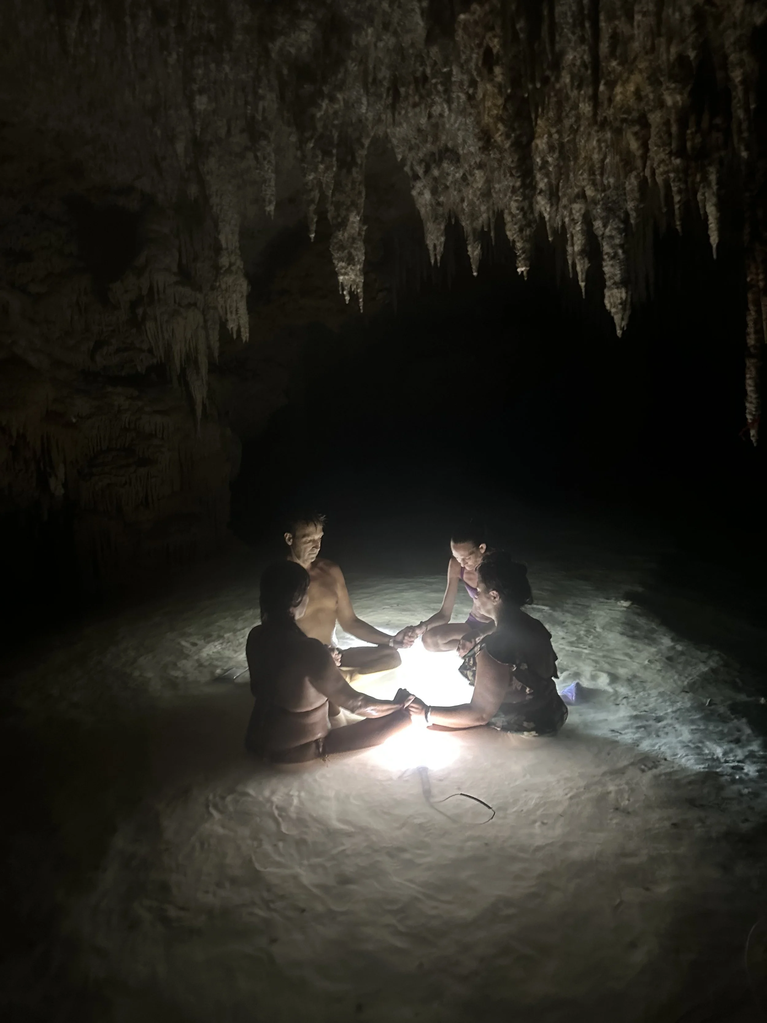 Four people sitting in a circle in a dark underground centoe cave, holding hands around a glowing light source on the sandy floor, with stalactites hanging from the ceiling.