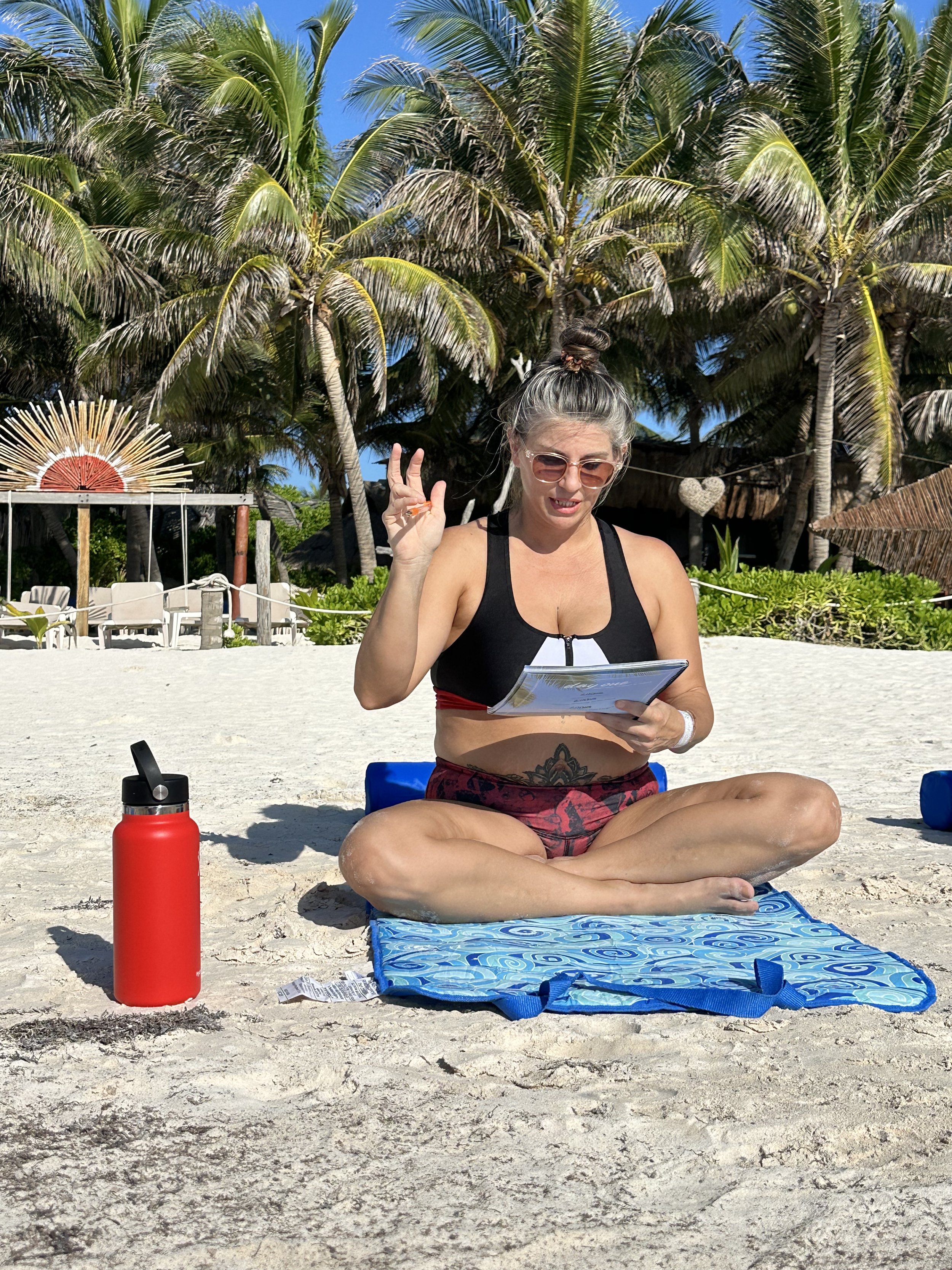 A woman sitting cross-legged on a blue mat on a sandy beach, holding a tablet and making an peace sign with her right hand, wearing sunglasses, a black sports top, and red shorts. There is a red water bottle to her left, with palm trees and beach huts in the background.