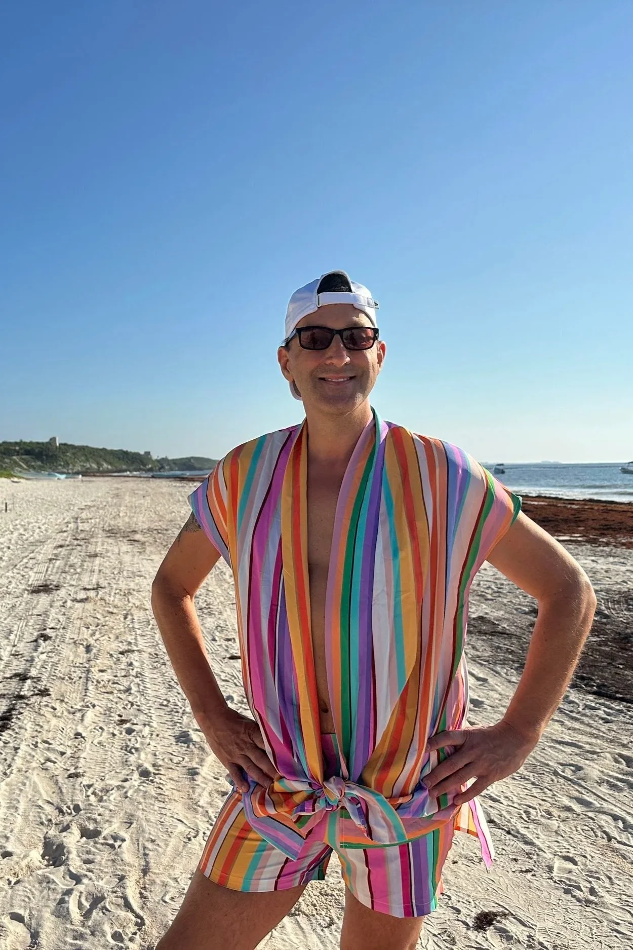 Man standing on beach with hands on hips, wearing rainbow-striped shirt and shorts, white cap backwards, sunglasses, smiling, clear blue sky in the background.