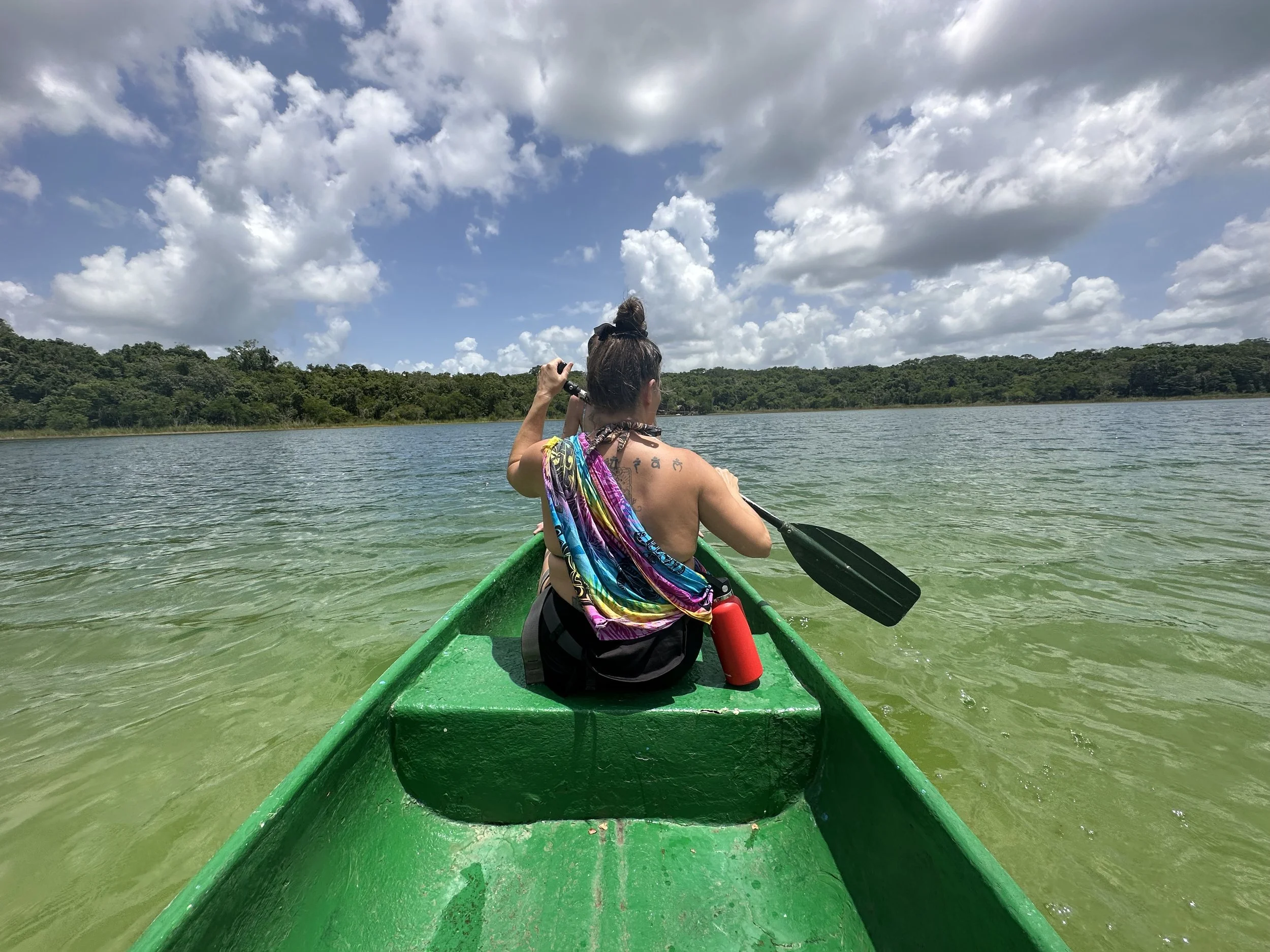 Person kayaking on a calm lake under a partly cloudy sky, surrounded by green trees in the distance.