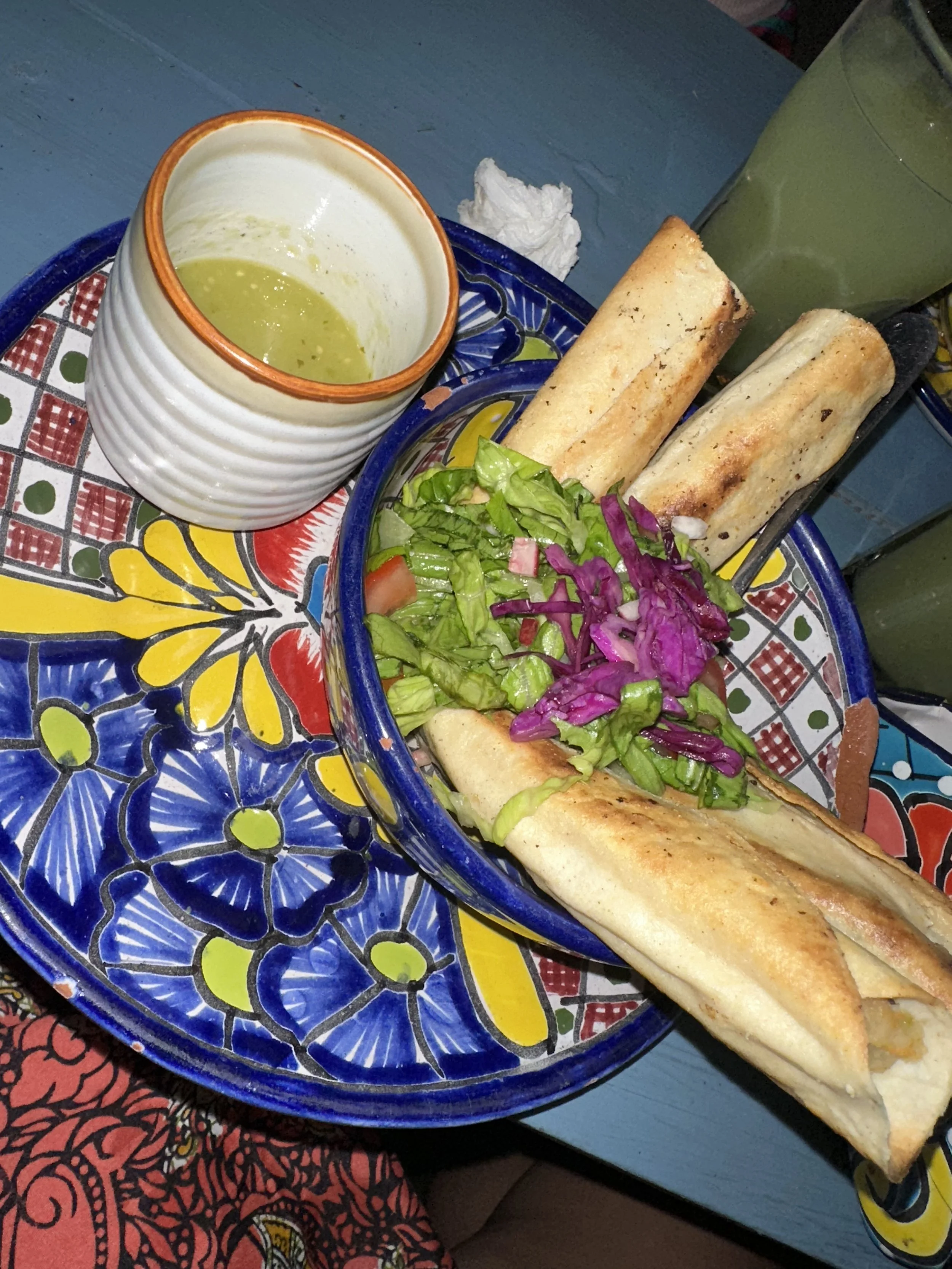 A colorful Mexican meal featuring two taquitos, a side salad with shredded lettuce and purple cabbage, a small bowl of green salsa, and two green drinks on a vibrant, patterned plate.