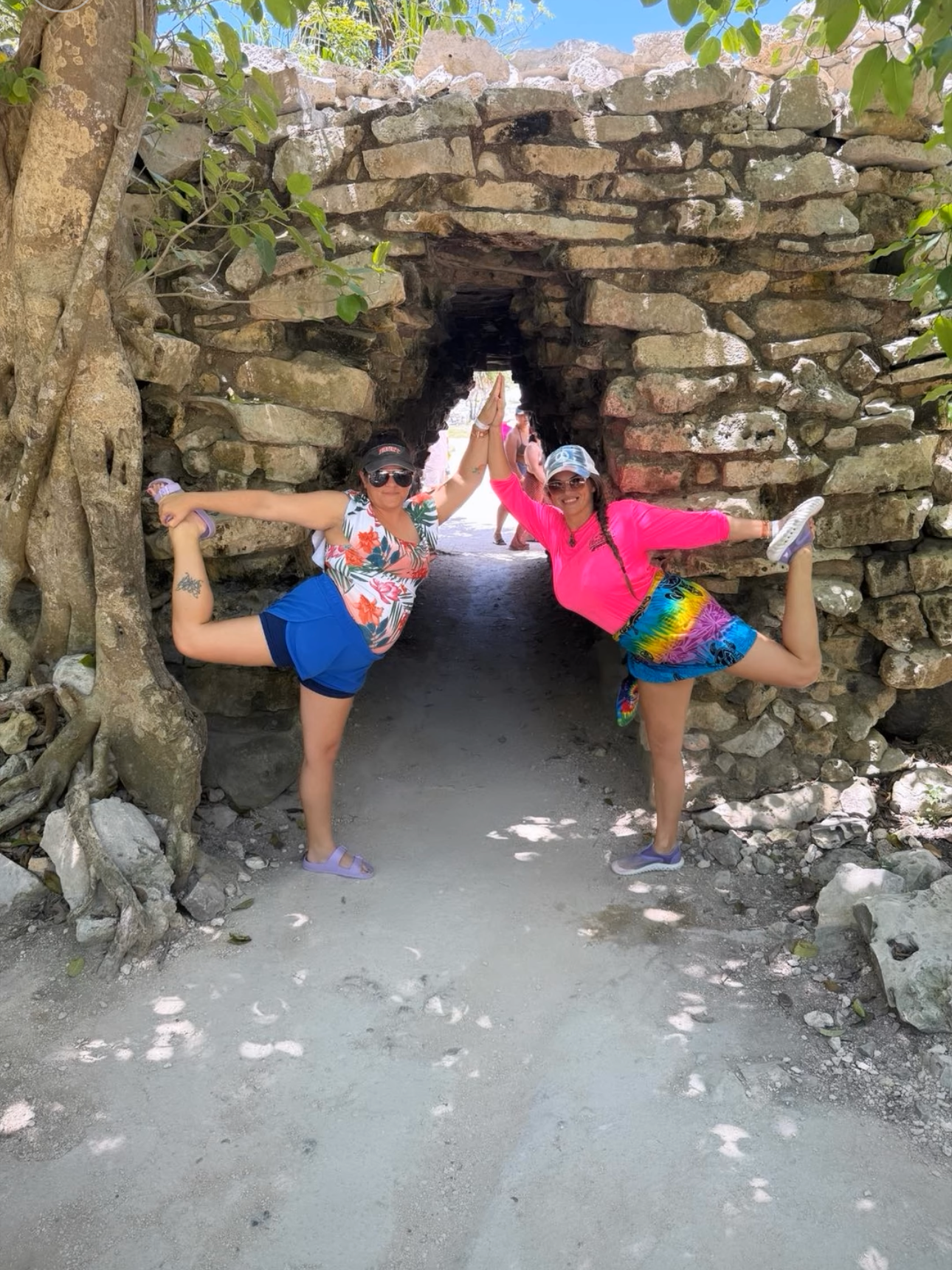Two women practicing yoga dancer pose in front of a stone archway under a tree, with a sunny sky and entering ancient ruins.