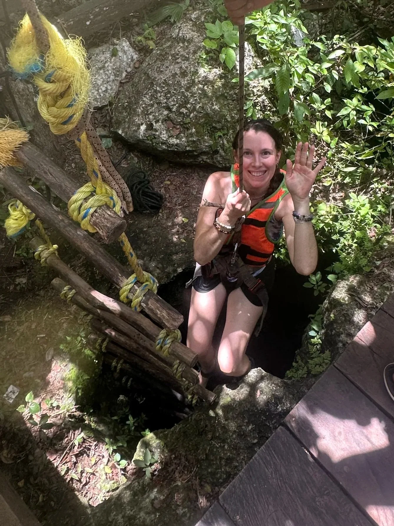 Woman dressed in outdoor gear, smiling and waving, standing in a hole surrounded by greenery descending into a centoe cave, with a wooden ladder and colorful ropes nearby in a forest setting.