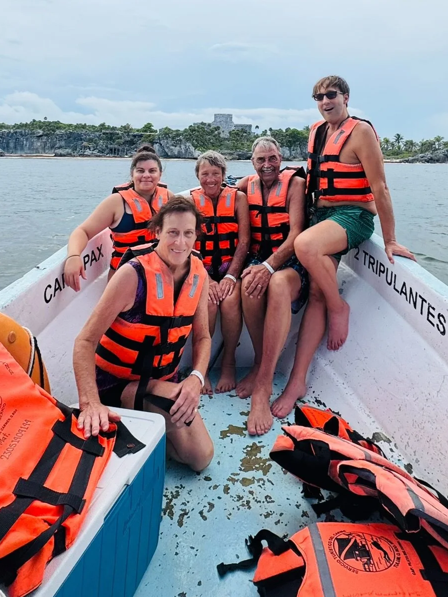 Five people wearing orange life jackets sitting and standing inside a boat on the water, with a rocky shoreline and trees in the background.