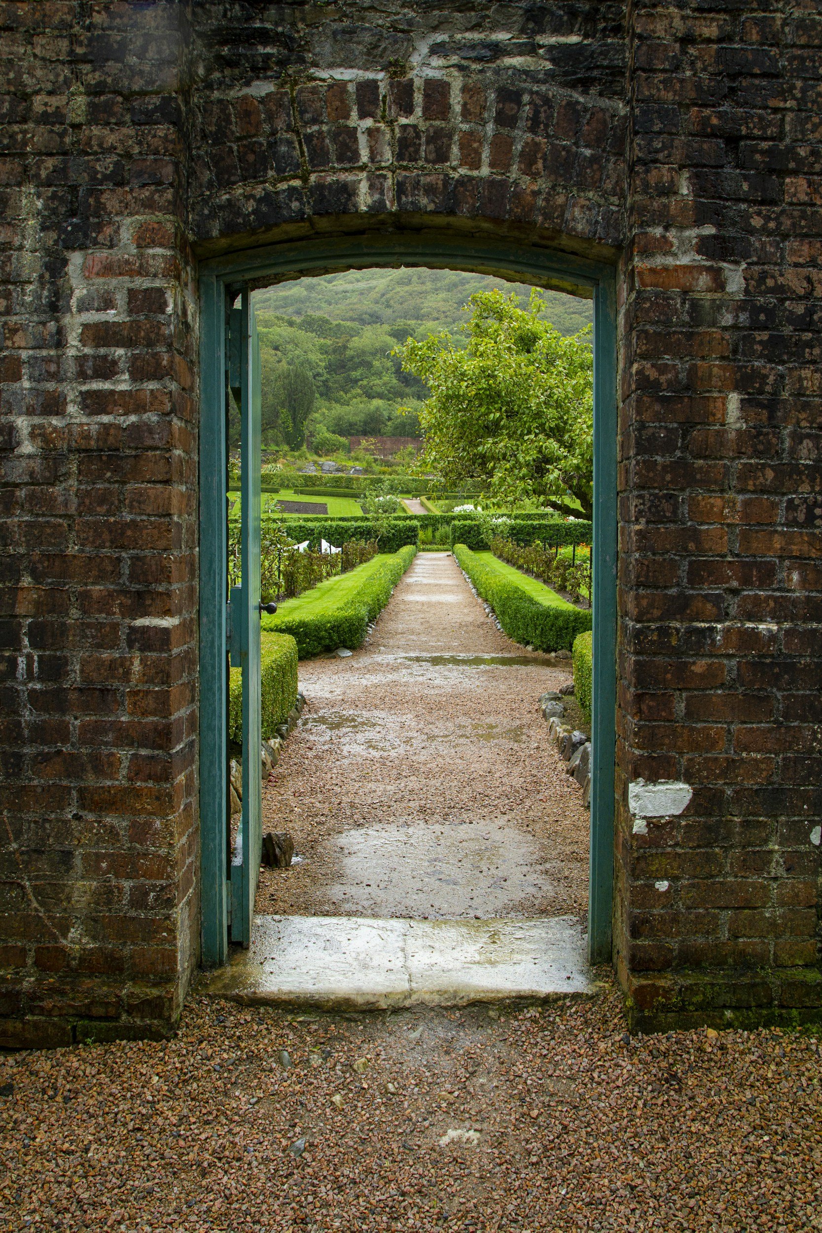 View of a garden path through a brick archway, with greenery and hills in the background.