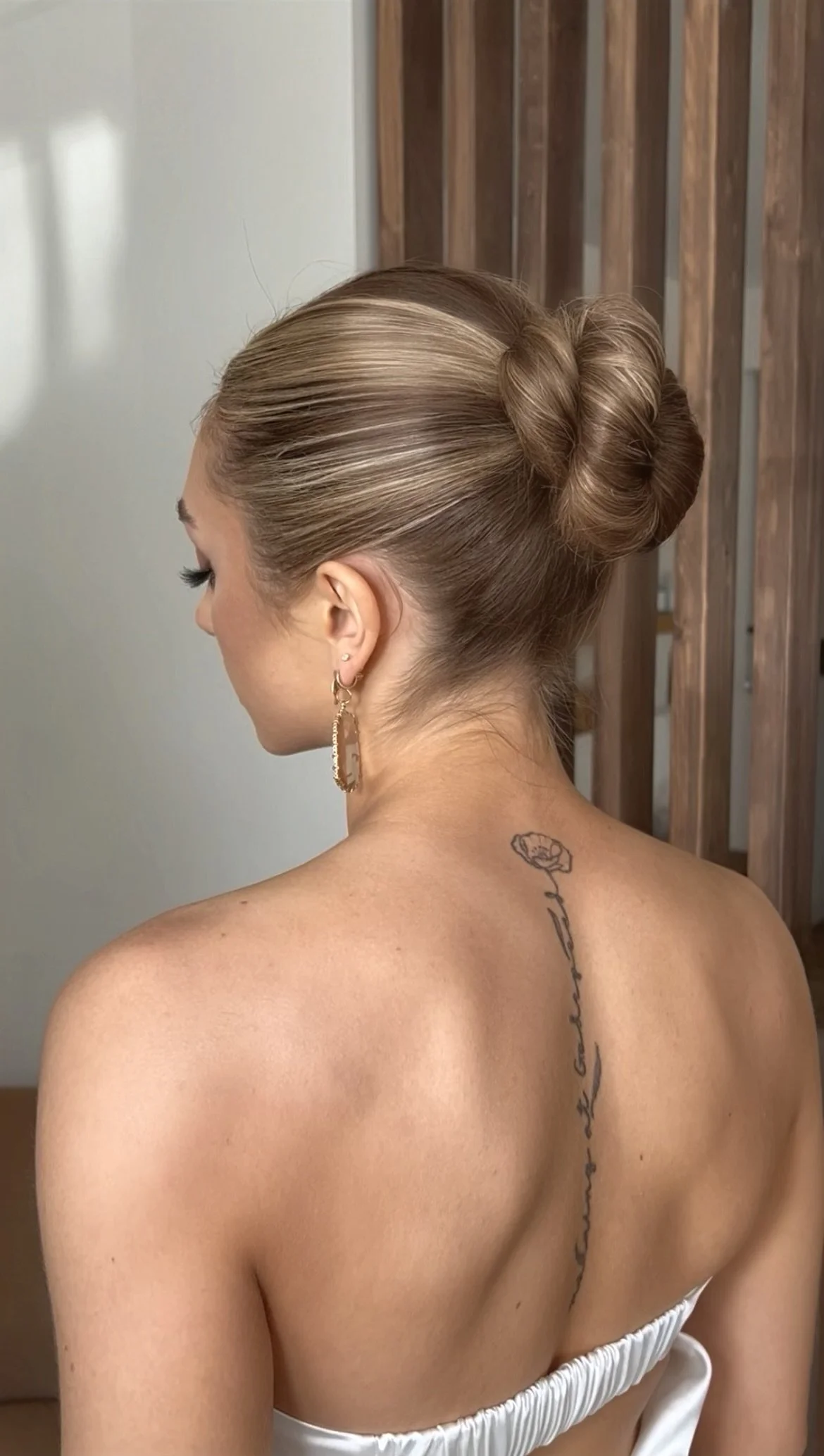 A woman with styled hair in a bun, wearing earrings, and a tattoo on her back, facing away from the camera.