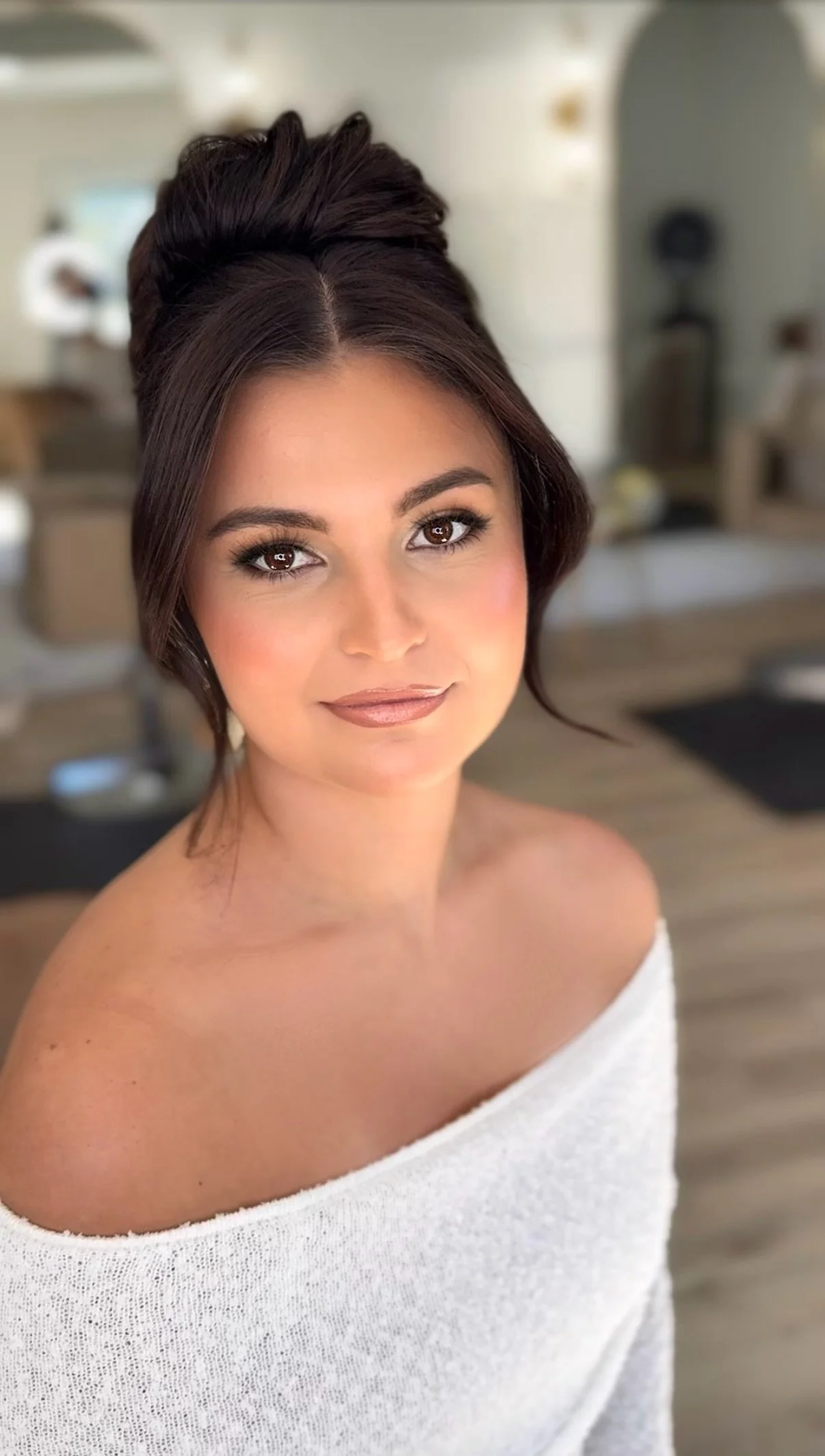 Close-up photo of a woman with styled brown hair, wearing a white off-shoulder top, smiling softly at the camera in a bright, modern room.