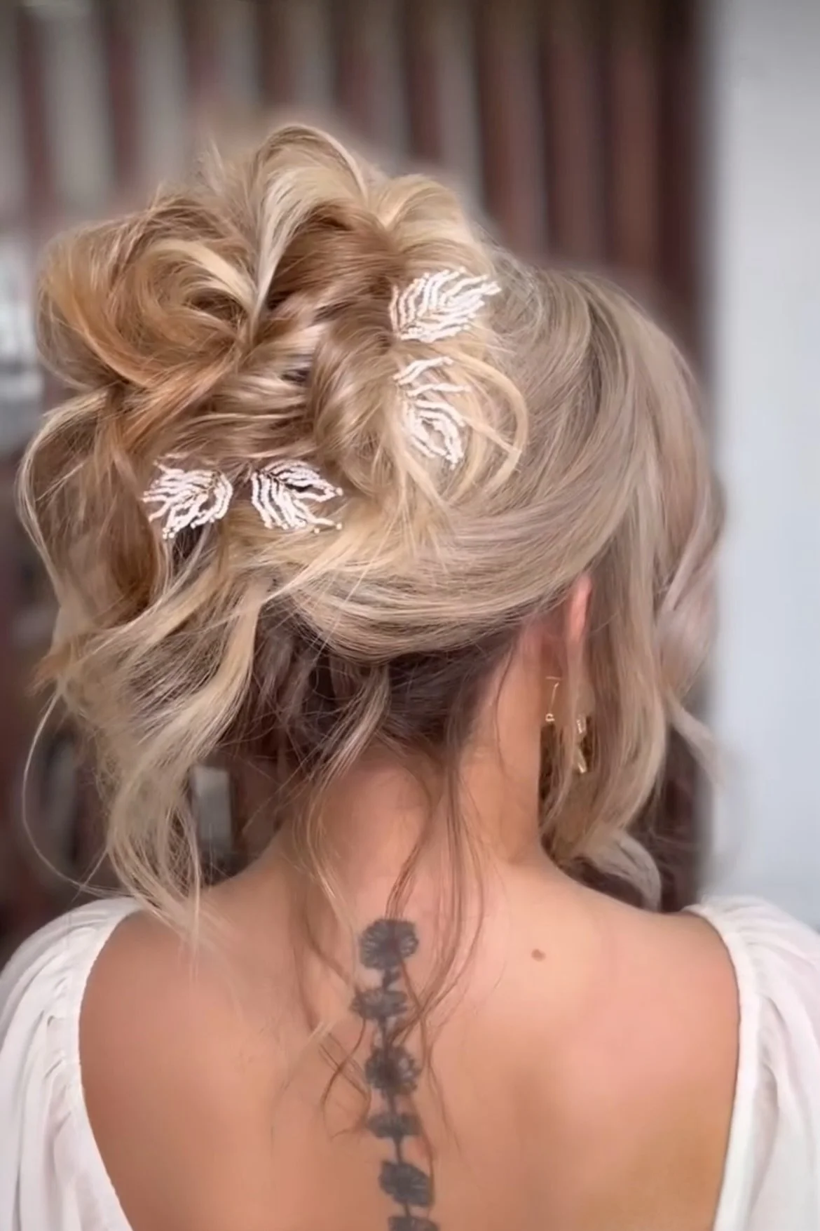 Back of a woman's head with blond, curled hair adorned with white leaf-shaped hair accessories. She has tattoos on her upper back and is wearing earrings, with a blurred background.