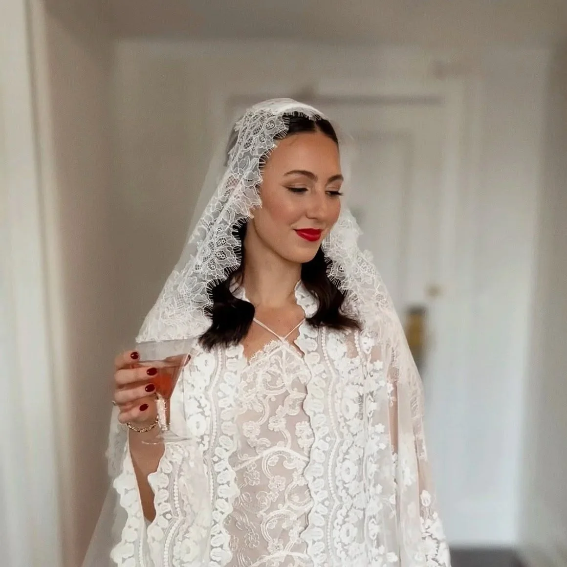 A woman in vintage wedding attire, wearing a lace veil and holding a martini glass, smiling softly.