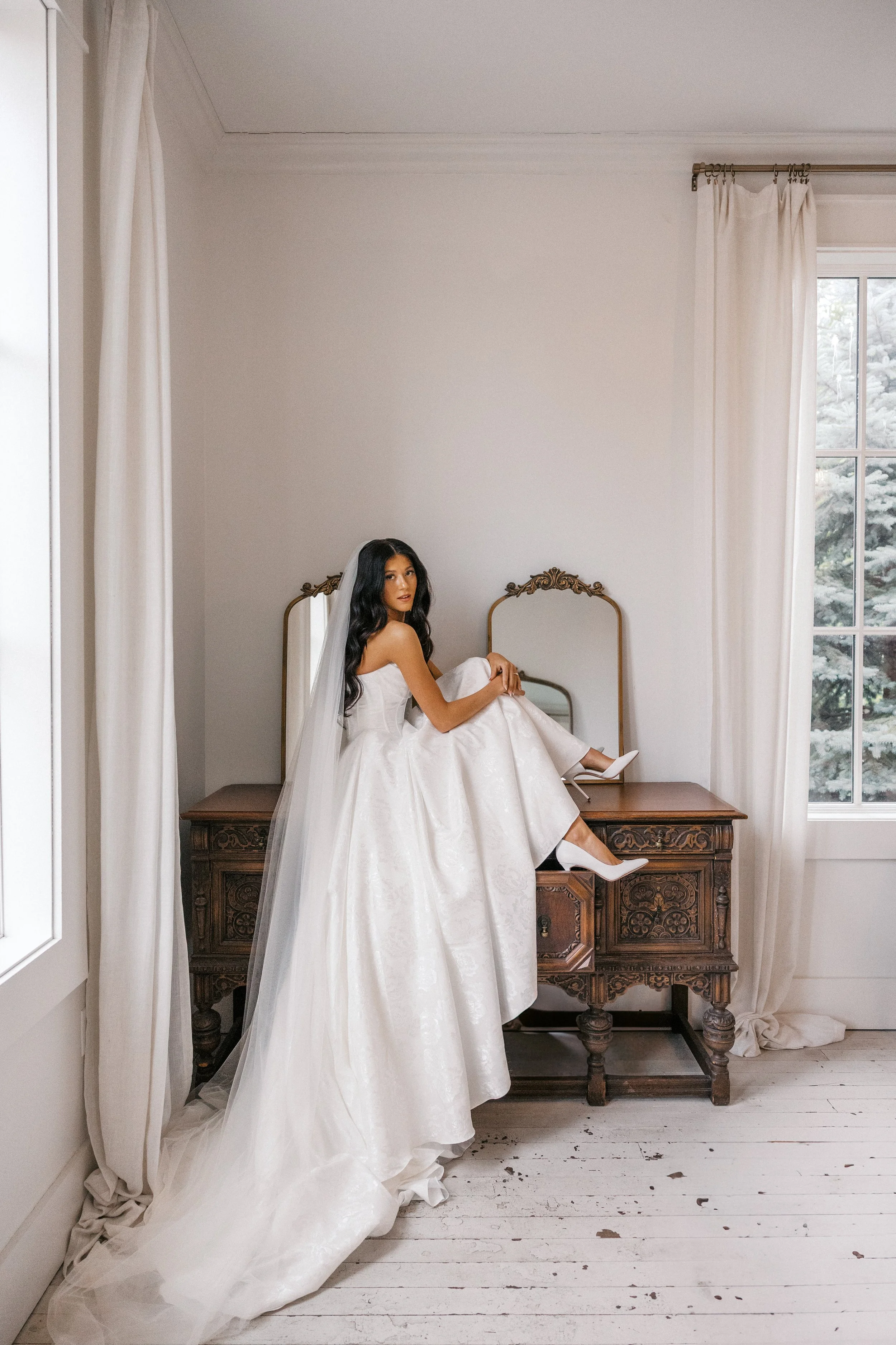 A bride in a white wedding dress and veil sitting on an antique wooden dresser with mirrors in a bright, minimalist room with white curtains and a window showing snow-covered trees outside.