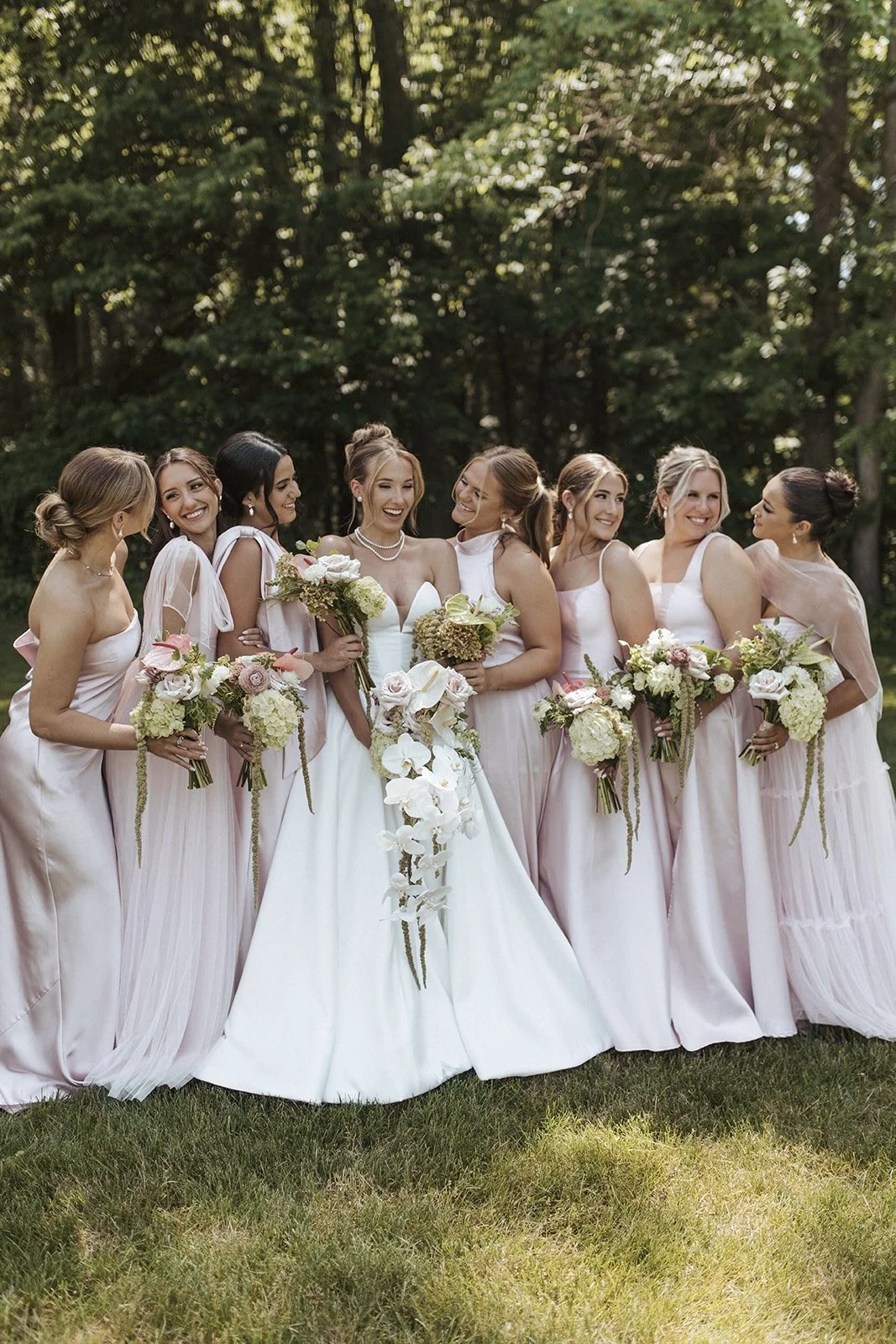 A bride surrounded by eight bridesmaids holding bouquets, standing outdoors on grass with trees in the background.