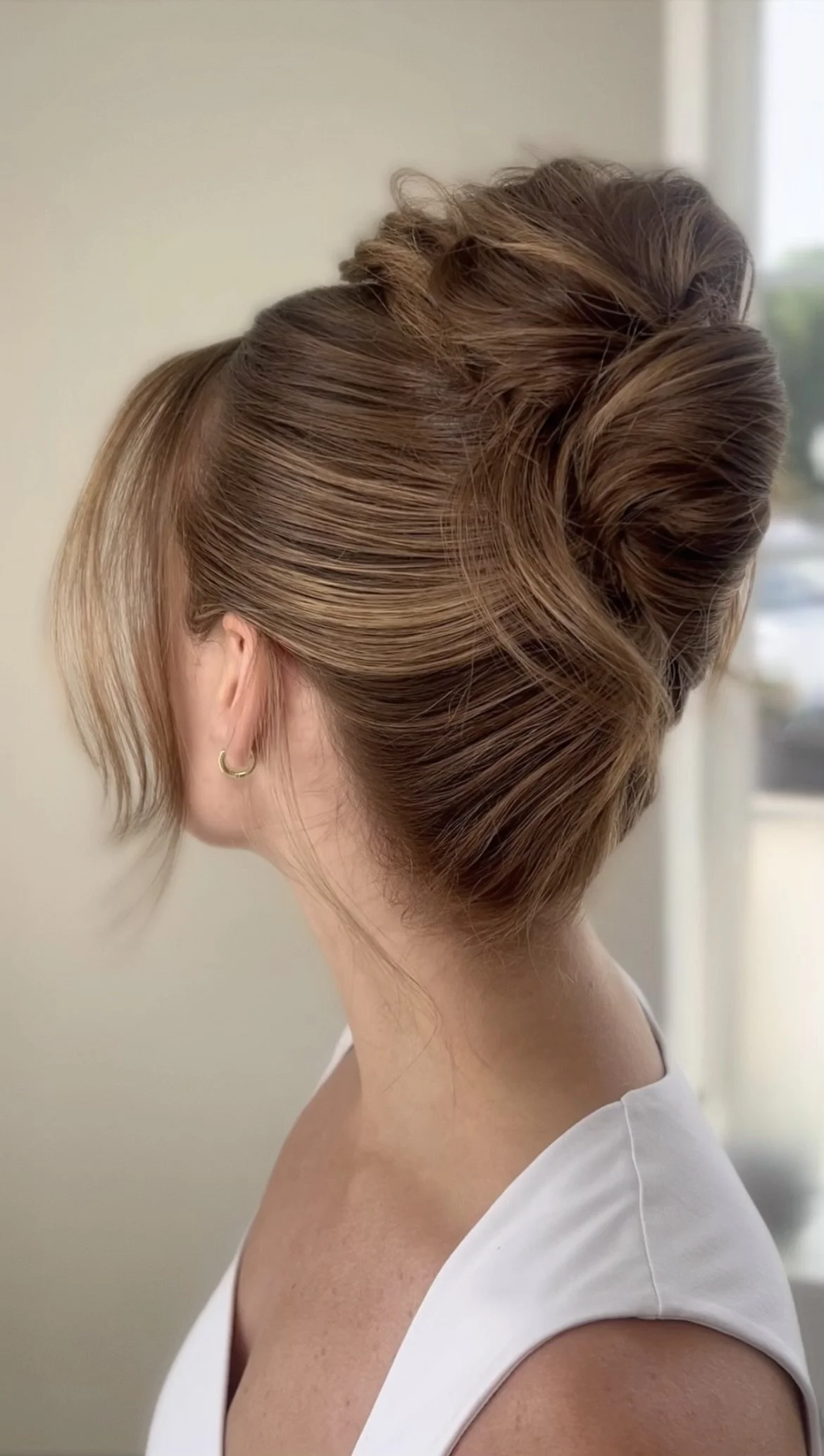 A woman with styled brown hair in an elegant updo, wearing a white sleeveless top and small gold hoop earrings, facing away from the camera.