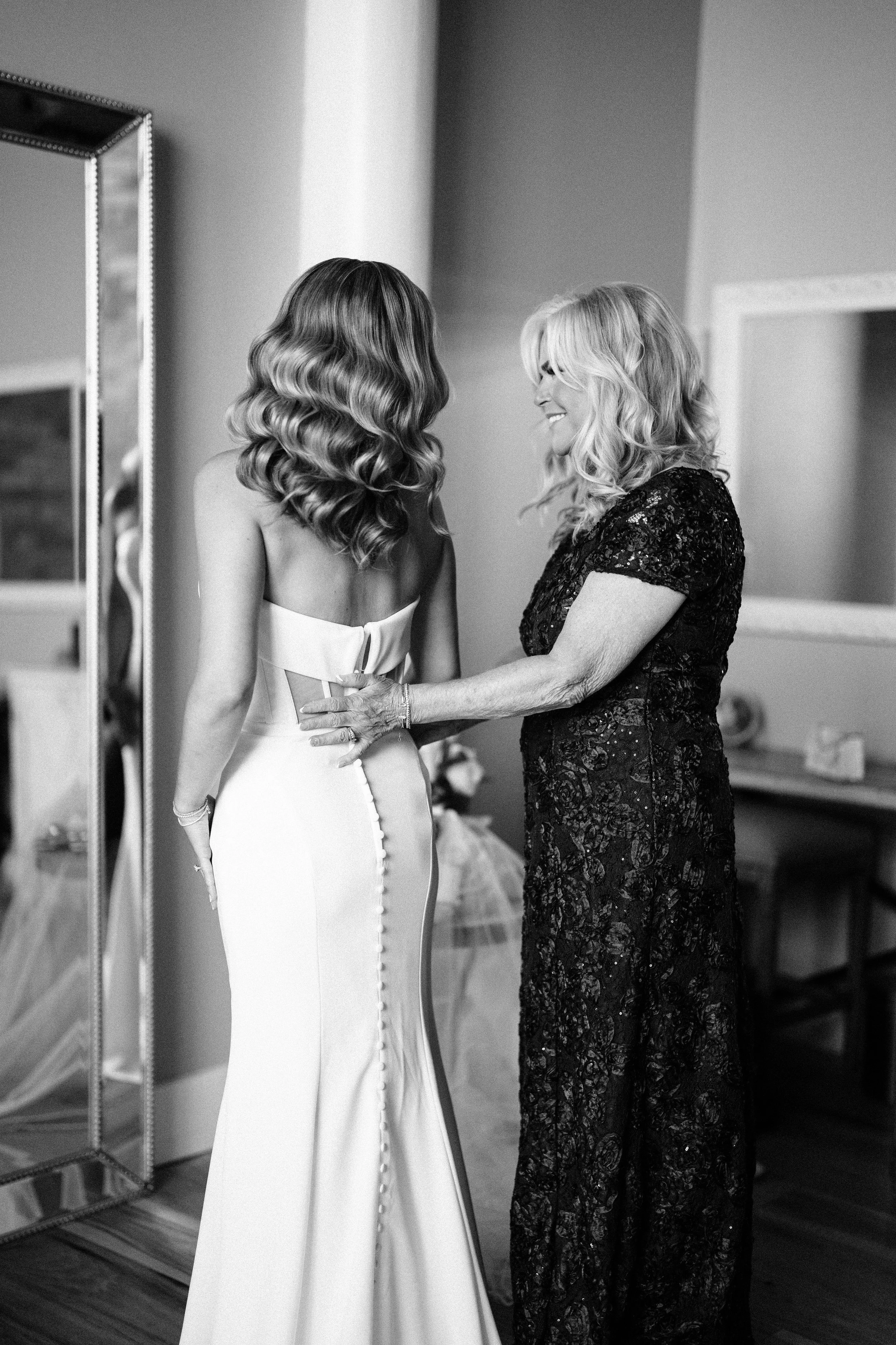 A bride in a strapless wedding dress with buttons down the back is smiling and embracing an older woman in a black lace dress, who is holding her waist.