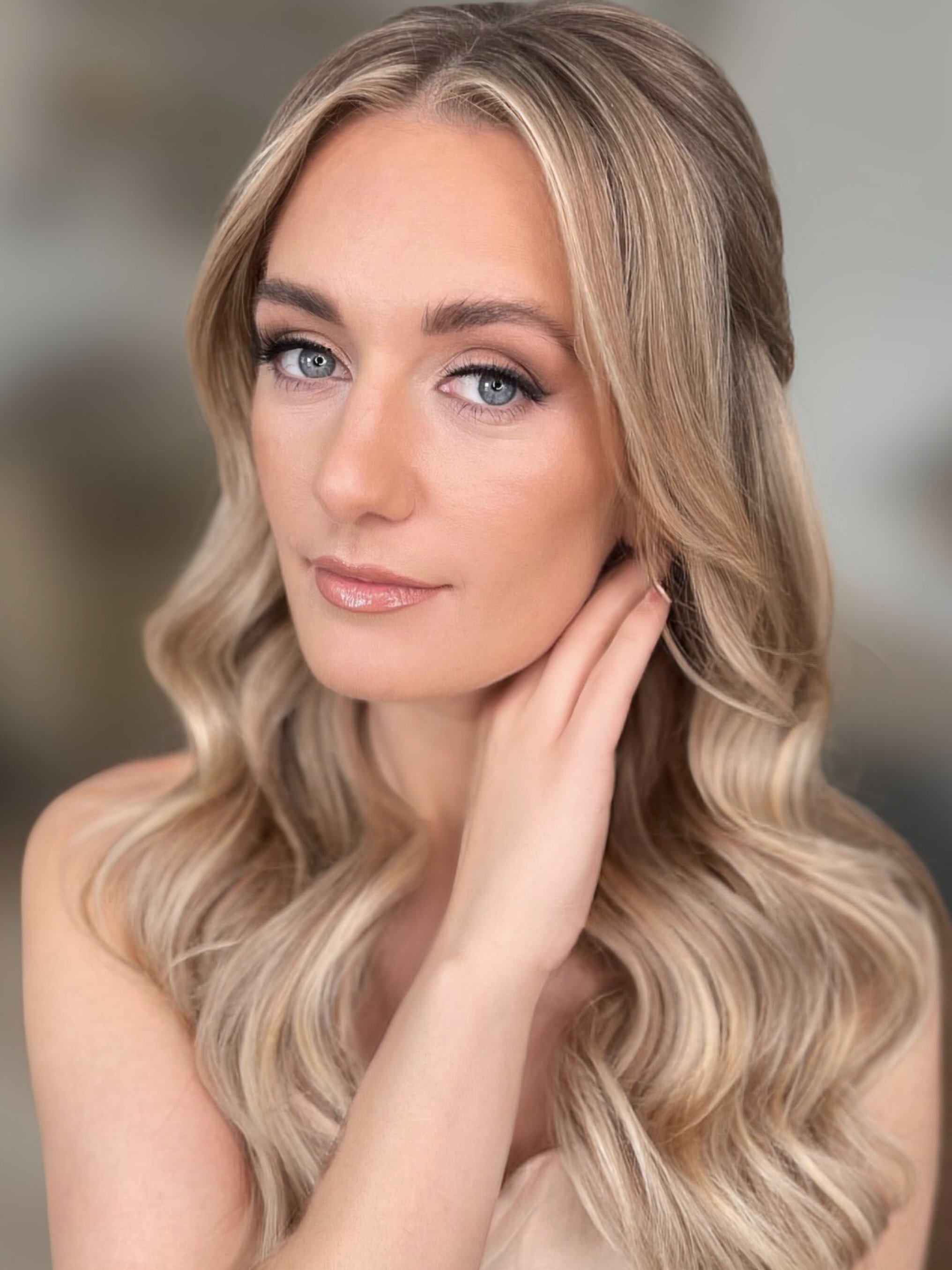 A woman with blonde hair styled in loose waves, blue eyes, and natural makeup, gently resting her hand on her face and looking at the camera.
