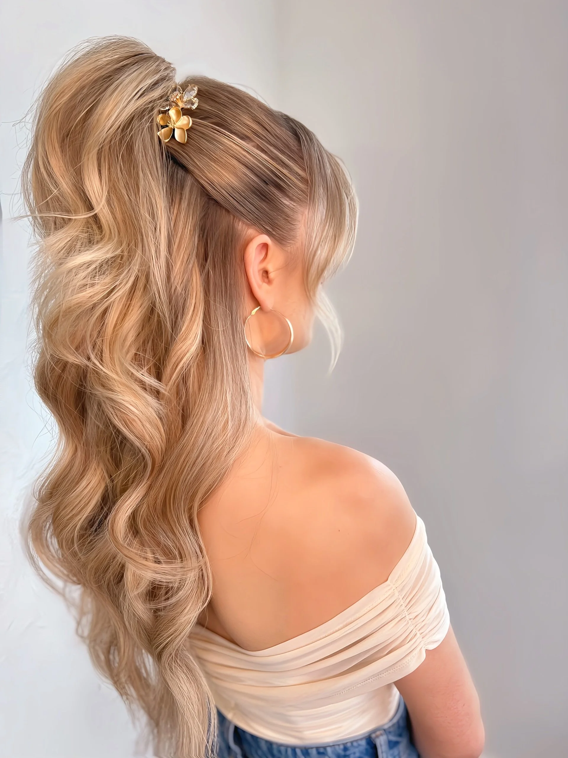 A woman with long, wavy blonde hair styled in loose curls, wearing a flower hair clip, large hoop earrings, an off-the-shoulder top, and sitting with her back to the camera against a plain background.