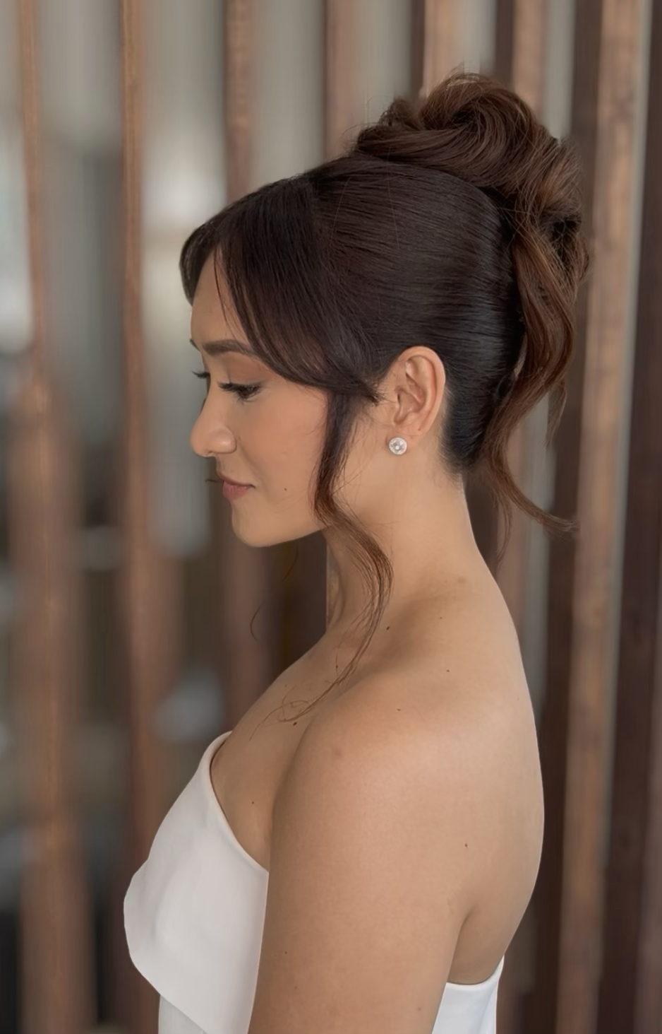 Side profile of a woman with dark brown hair styled in an elegant updo, wearing diamond stud earrings and a strapless white dress, standing in front of a wooden background.