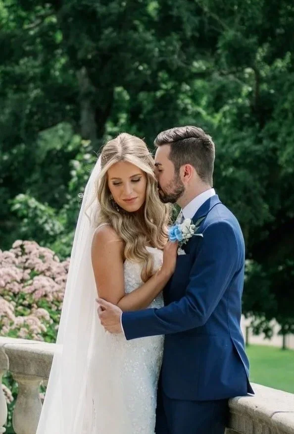 A bride and groom embracing outdoors during their wedding, with lush green trees and blooming flowers in the background.